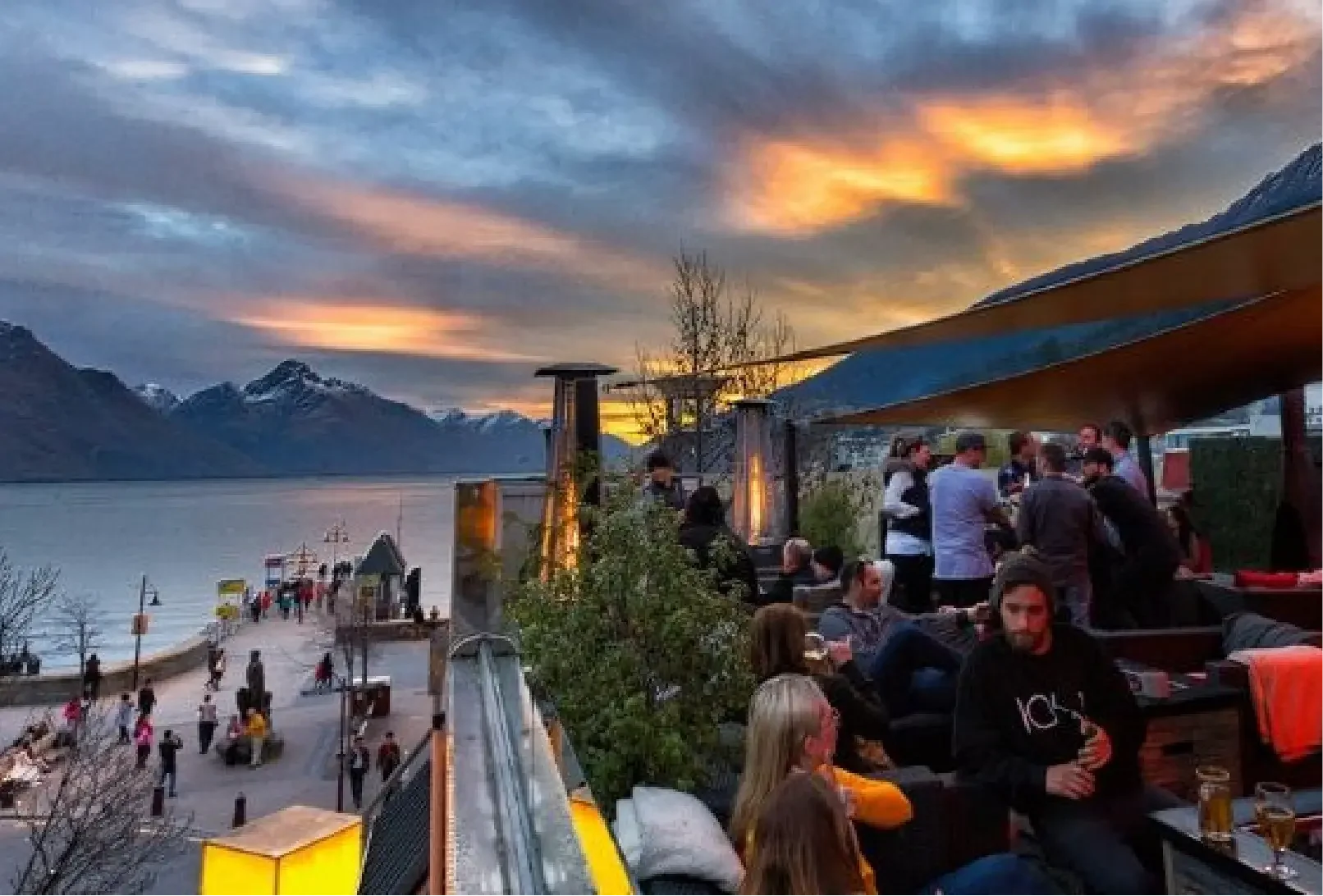 People gathering at an outdoor lakeside restaurant during sunset, with mountains in the background.