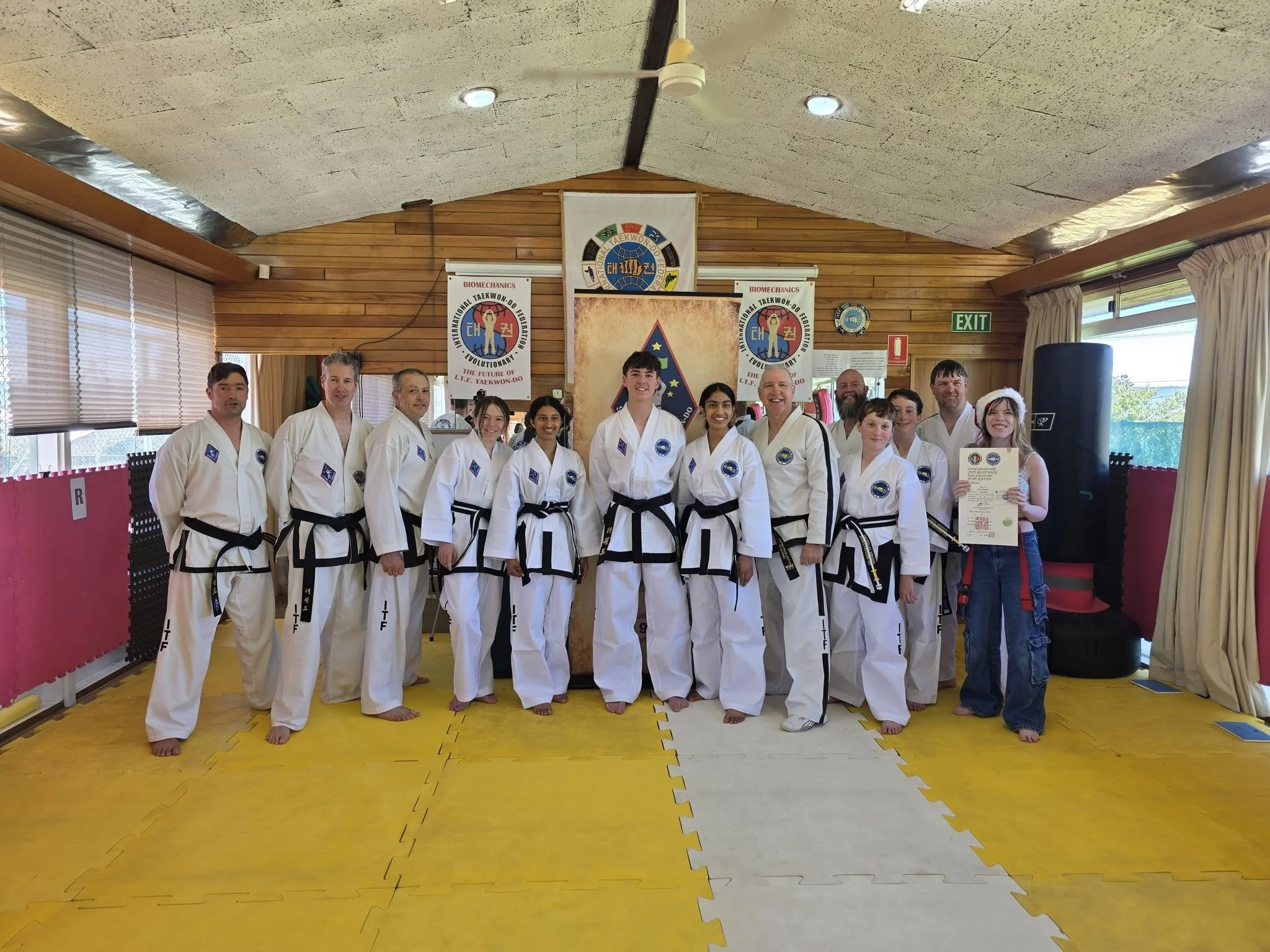 Group of martial artists in white uniforms and black belts standing in a dojo, posing for a group photo with patches on their uniforms, in front of banners and a flag.