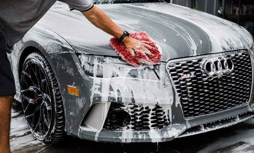 Person washing a silver Audi car with a red sponge and soap suds.
