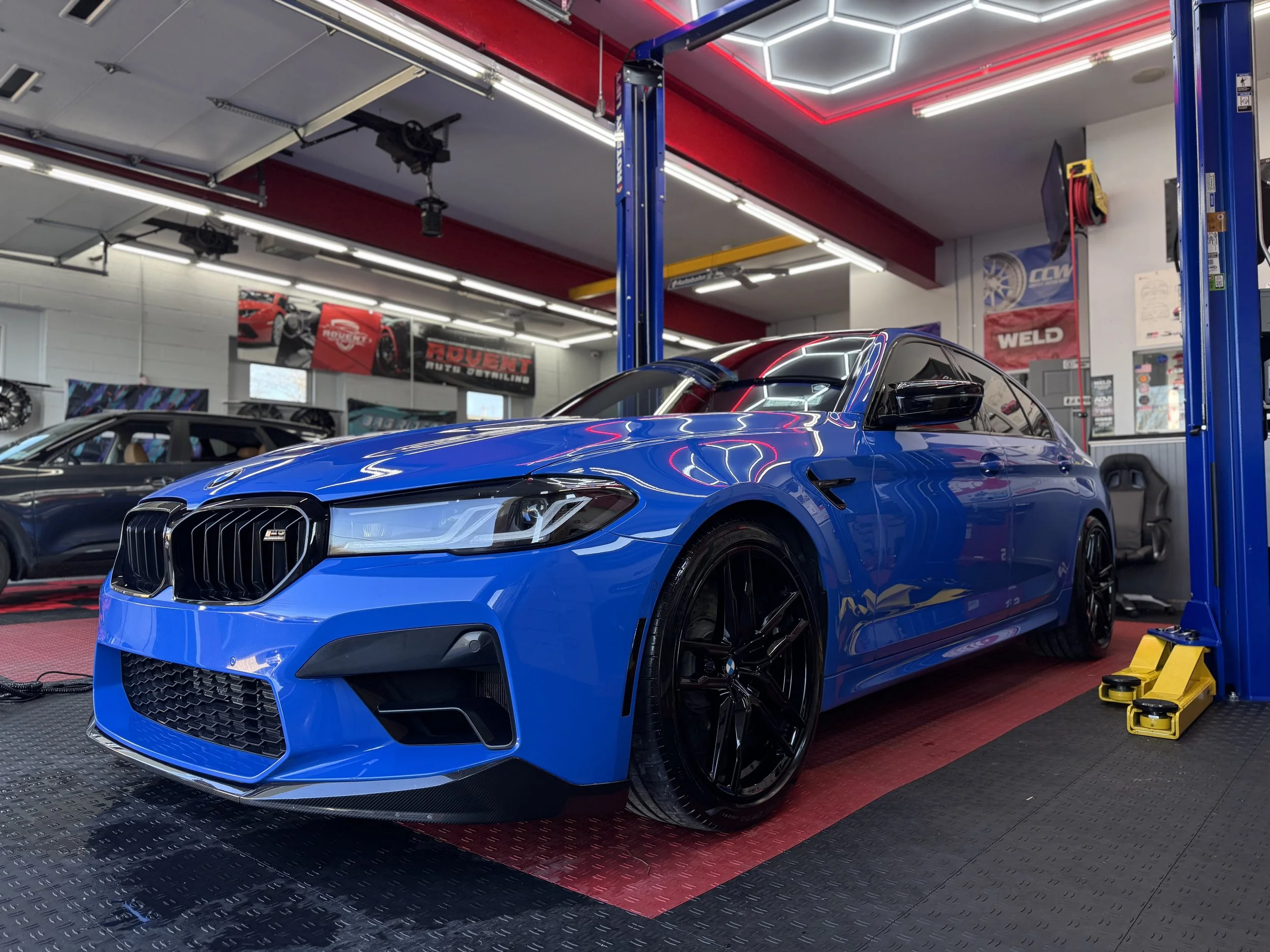 A blue BMW sedan lifted on a hydraulic car lift in an automotive workshop, with a black roof and black wheels, surrounded by automotive equipment and posters.