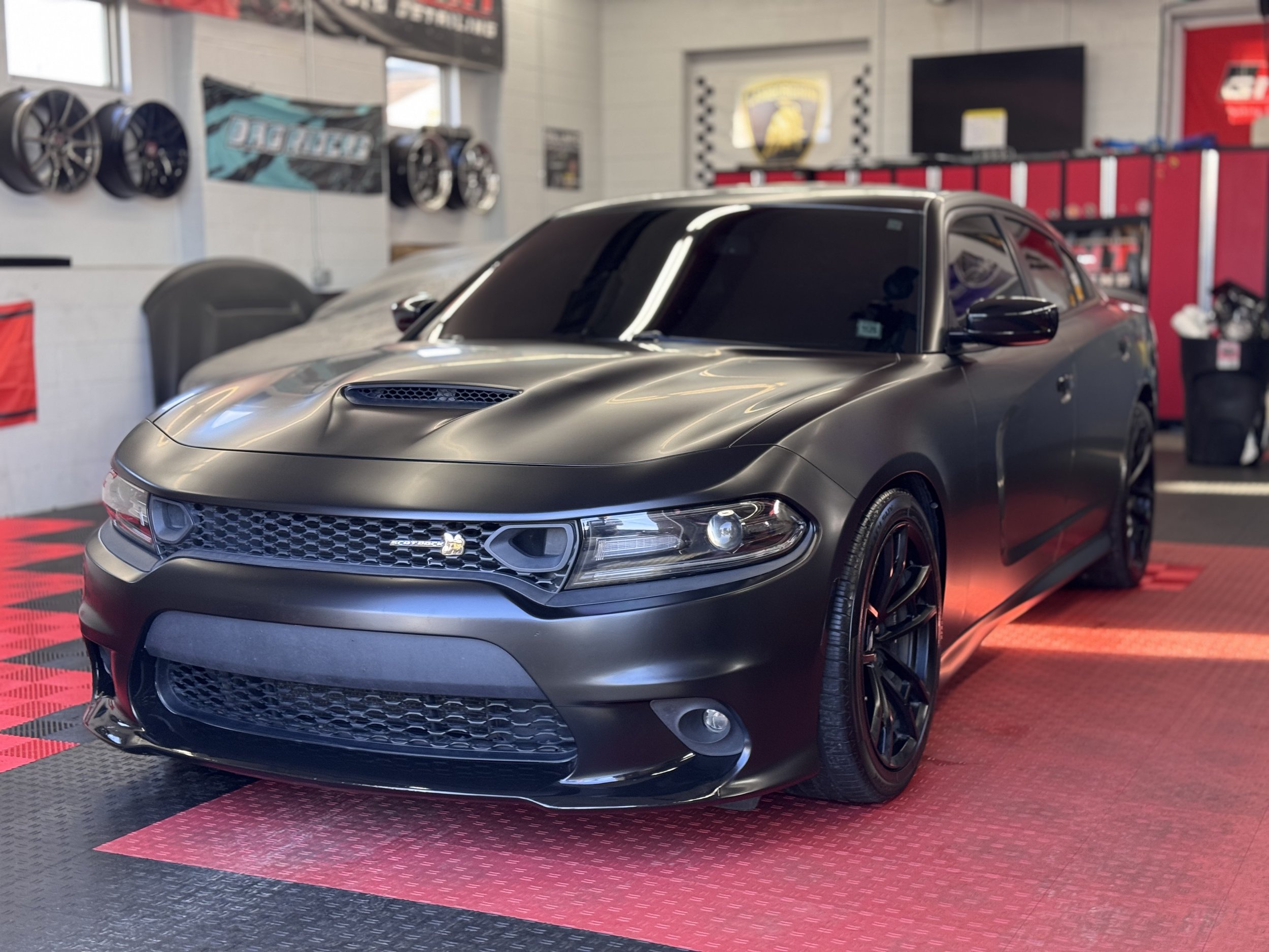 A black Dodge Charger sports car on display in a garage with red and black flooring, surrounded by wheels and automotive supplies.