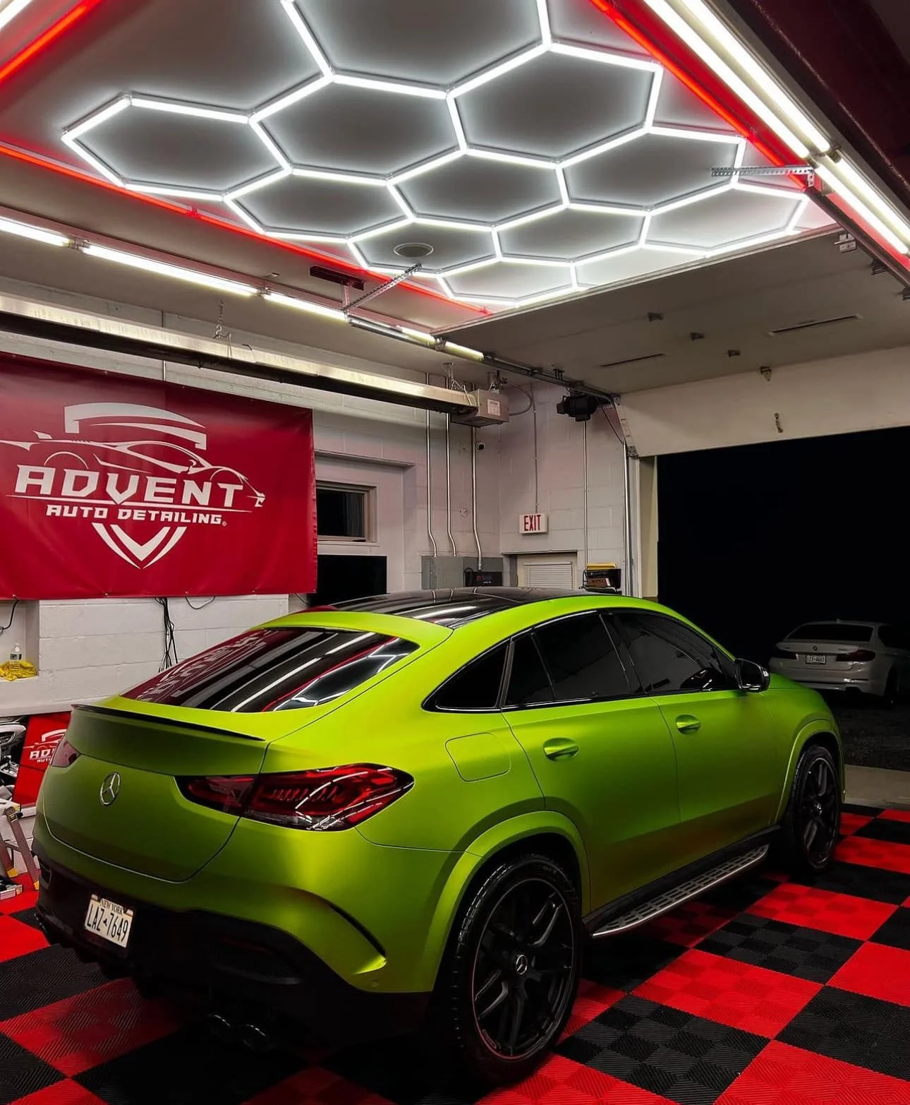 A bright green Mercedes-Benz SUV with tinted windows parked inside a car detailing garage. The garage has hexagonal LED ceiling lights and a red and black checkered floor. A red banner on the wall reads 'Advent Auto Detailing'.