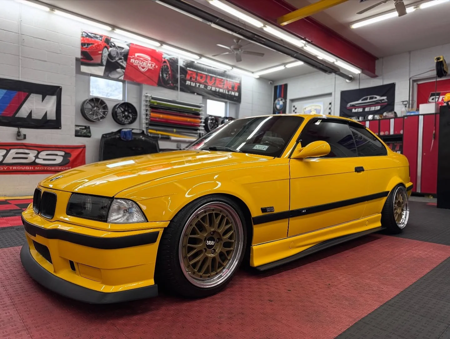A yellow BMW E31 M3 with aftermarket wheels inside a garage, surrounded by various car banners, wheels, and tools.