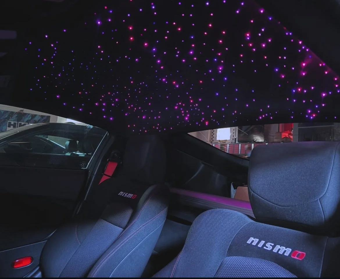 Car interior with a ceiling decorated with purple and pink star-like LED lights, two black seats with 'Nismo' embroidery, and a window showing a city street outside.