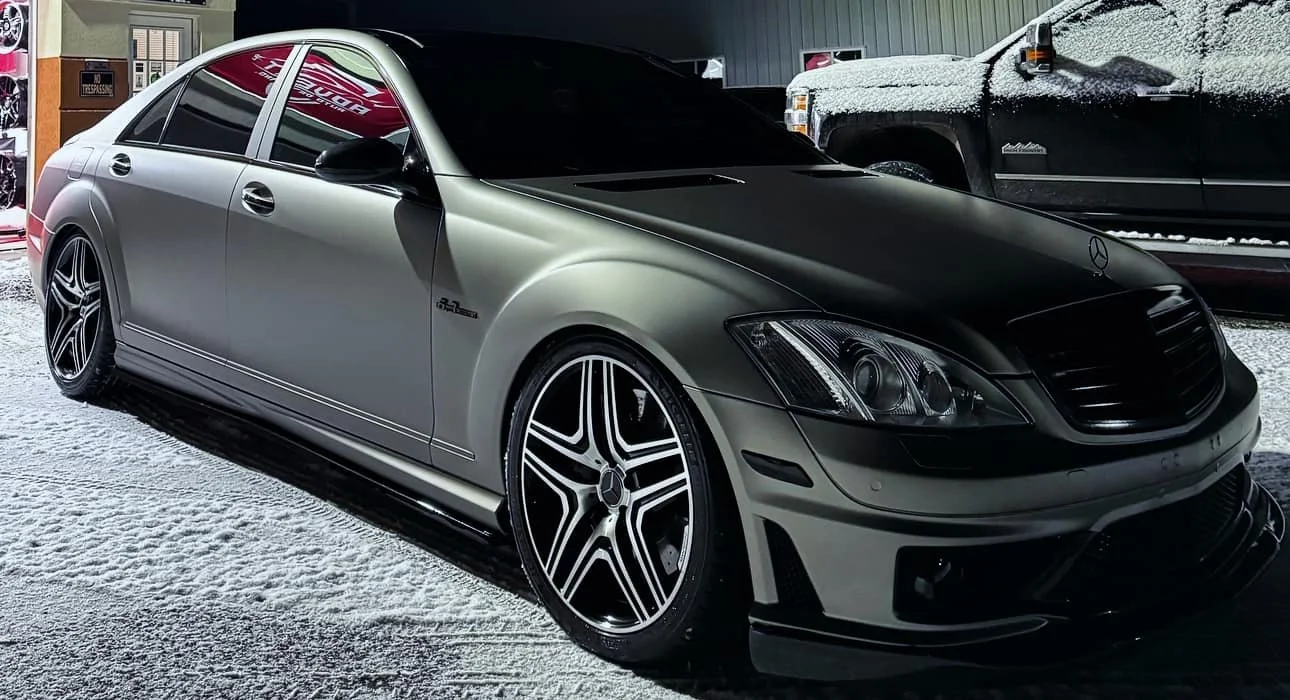 A silver Mercedes-Benz luxury sedan with a matte black hood, parked on a snowy surface, next to a black truck also covered in snow.