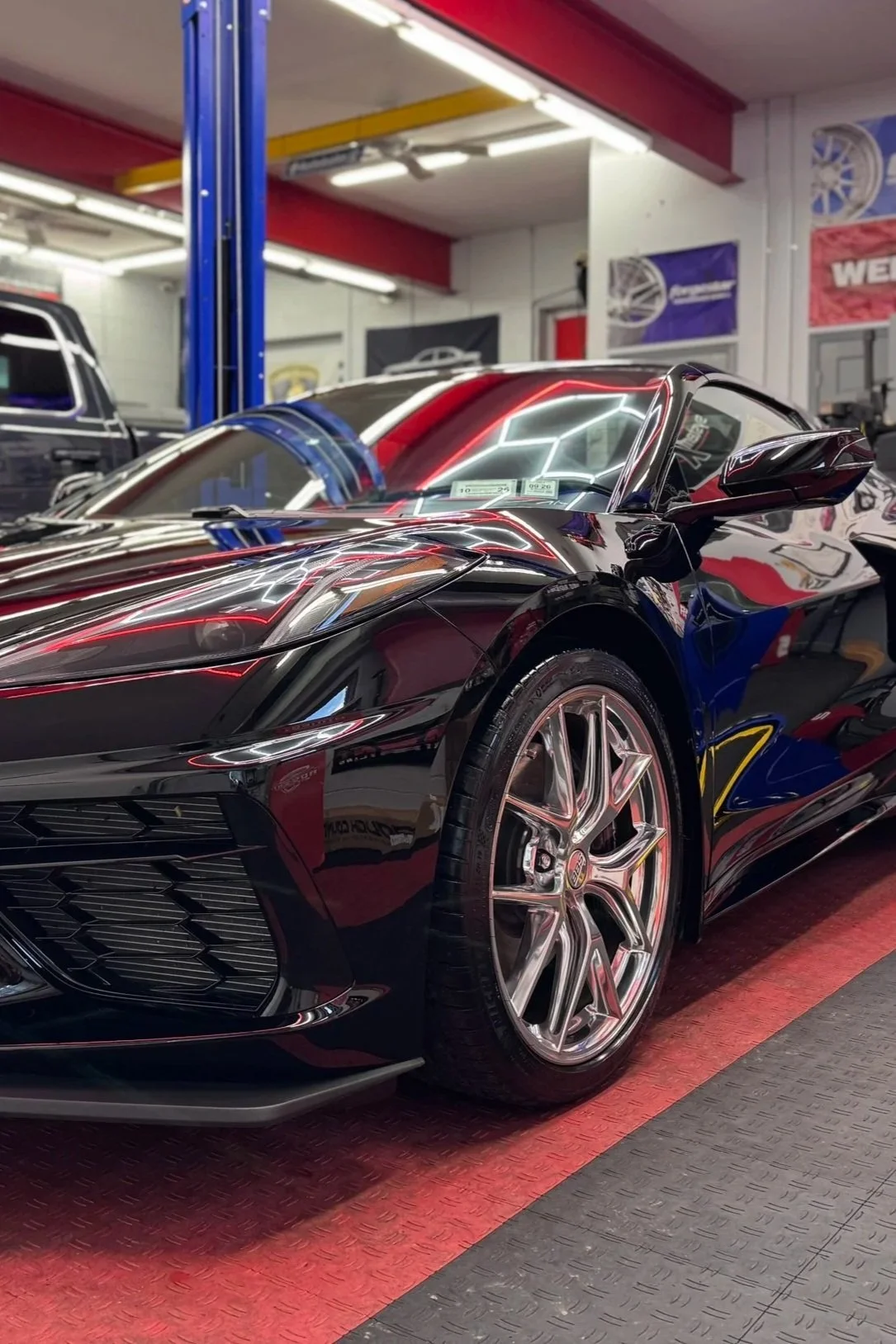 A shiny black sports car inside a garage or showroom, with reflections of bright lights and colorful banners on its surface.