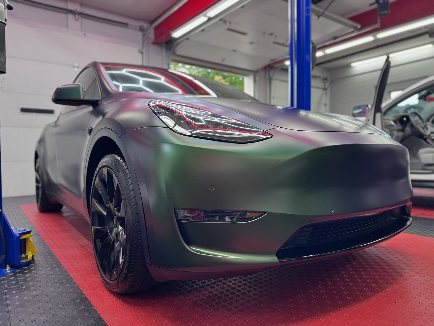 A sports car with a matte green finish parked in a garage, with a raised garage door and another vehicle visible in the background.
