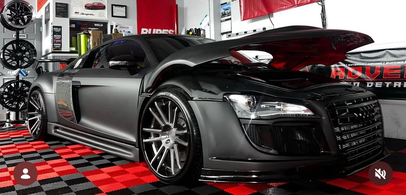 A silver and black Audi R8 sports car in a garage, with a raised rear hood showing the engine, surrounded by car wheels, tools, and automotive posters.