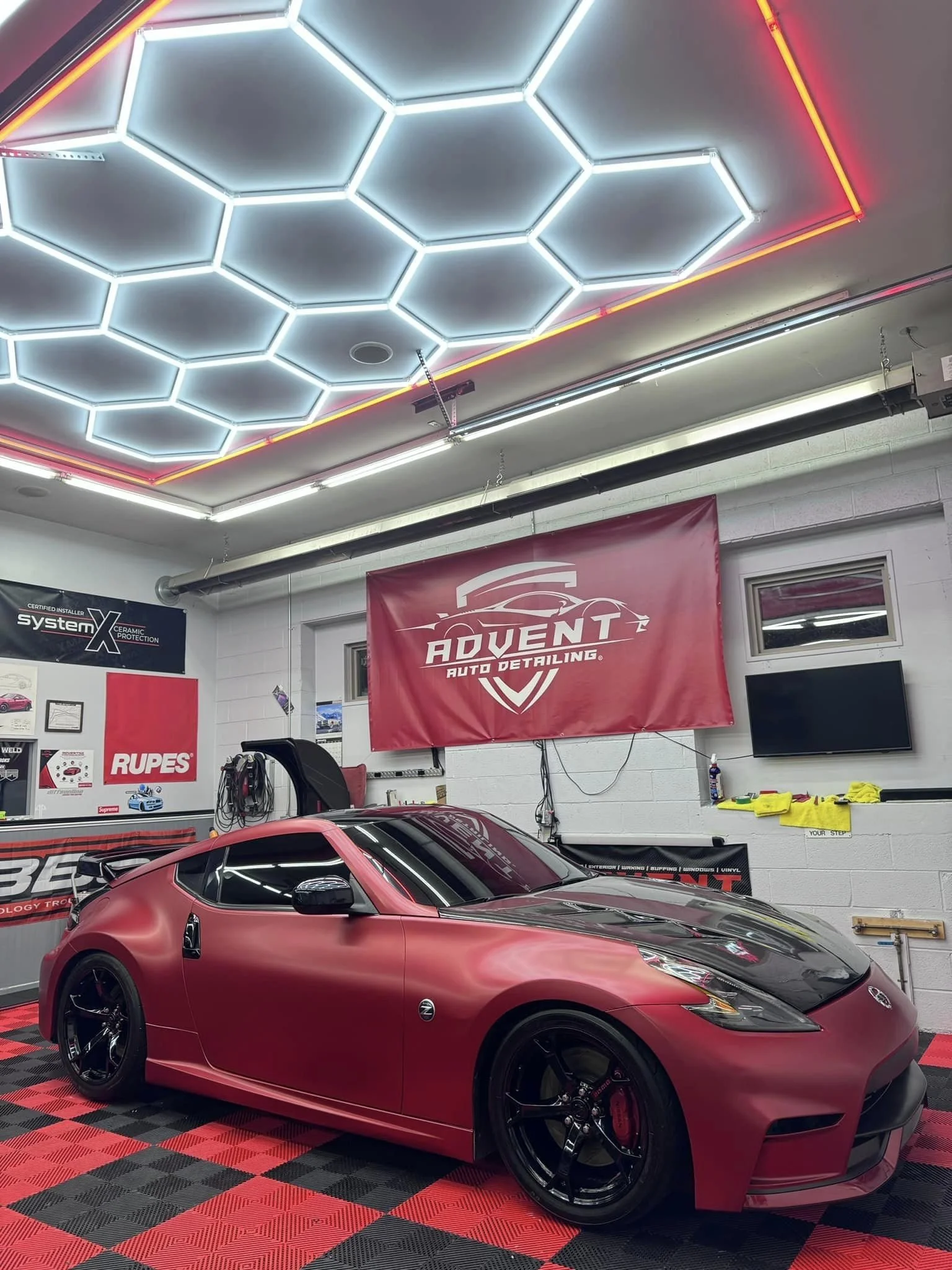 Red sports car in a professional auto detailing garage with neon hexagon ceiling lights, tools, and a large banner reading 'Advent Auto Detailing'.