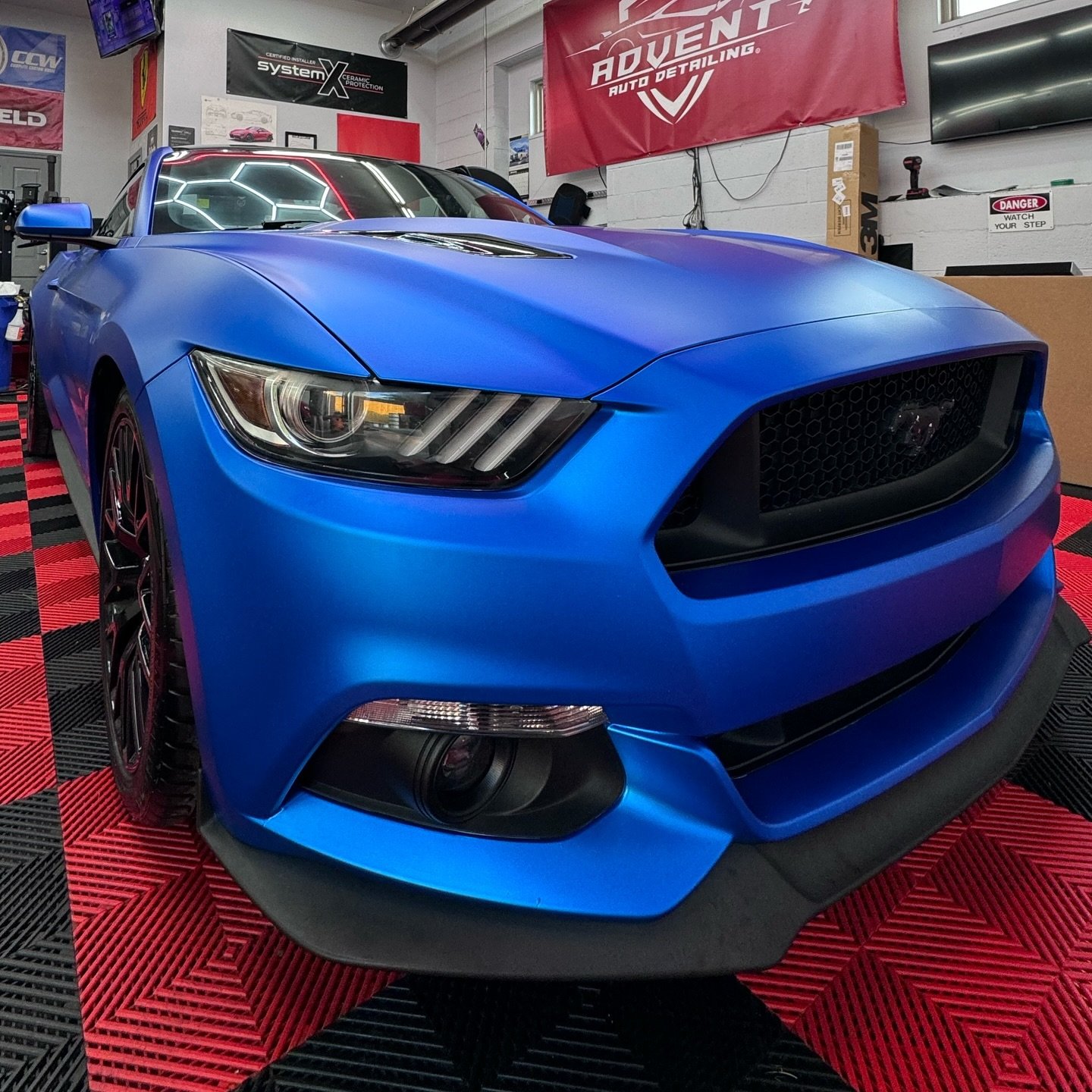 Blue Ford Mustang car inside an auto detailing shop with red and black checkered flooring, banners and equipment in the background.