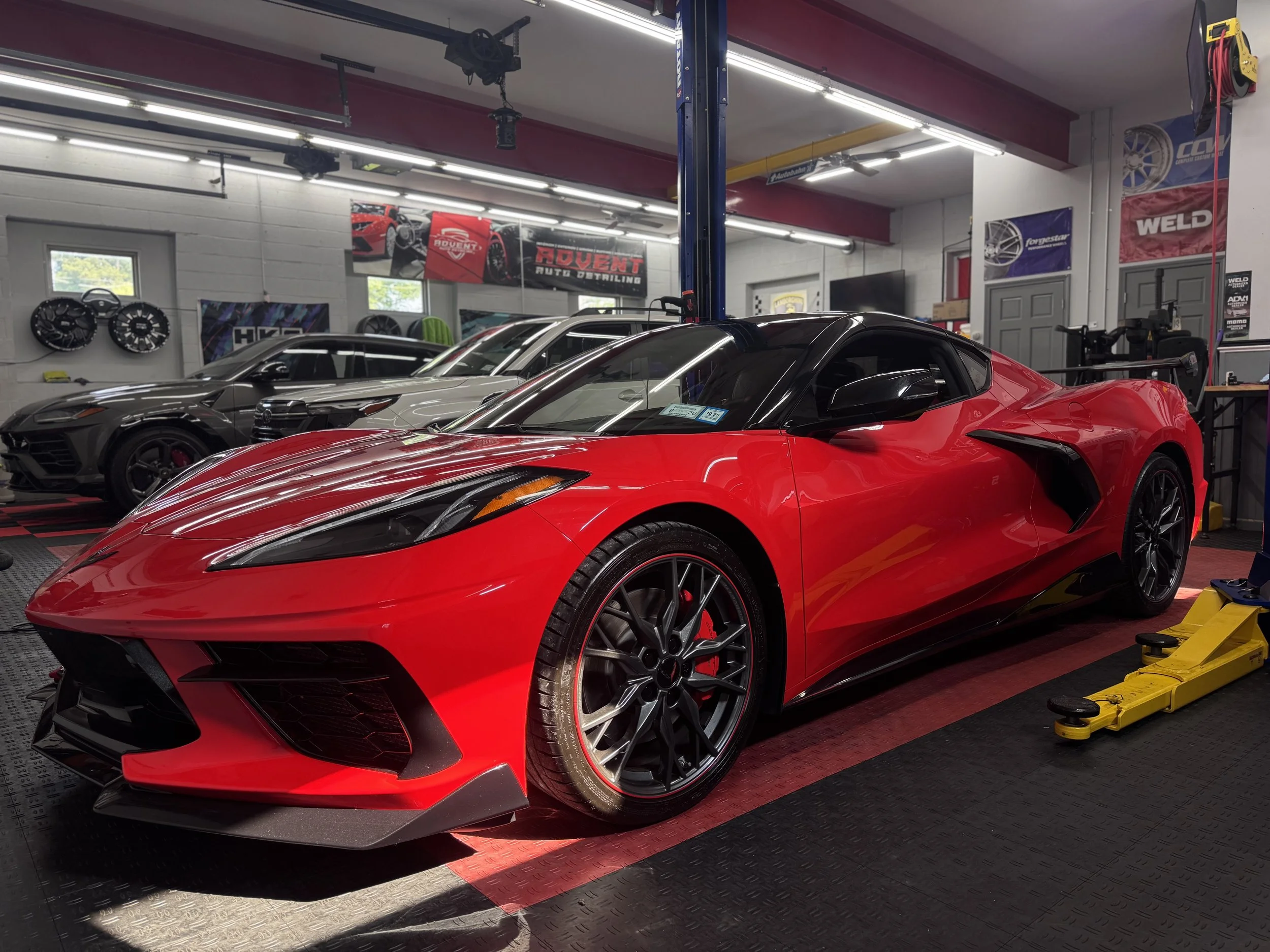 A red sports car parked inside a garage with other cars in the background and car wheel rims on the wall.