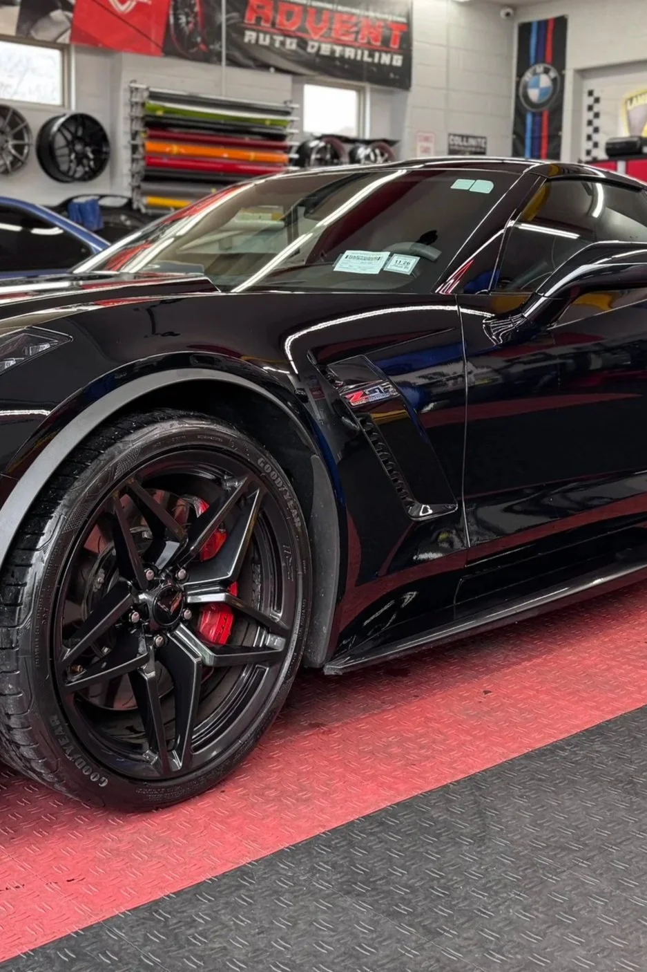 A black Chevrolet Corvette Z06 sports car inside an automotive workshop or detailing shop with various tools, wheels, and car wraps in the background.