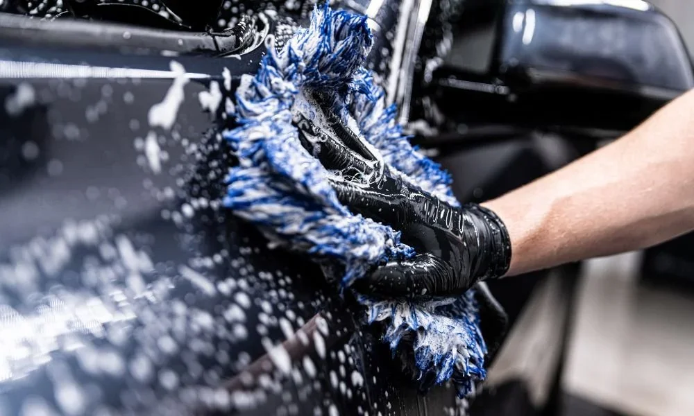 A person washing a black car with a blue and white microfiber cloth, using soap and water.