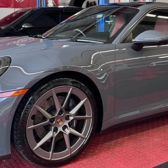 Close-up of a gray Porsche sports car, focusing on the front left wheel, side mirror, and door handle, parked on a red floor in an indoor showroom.