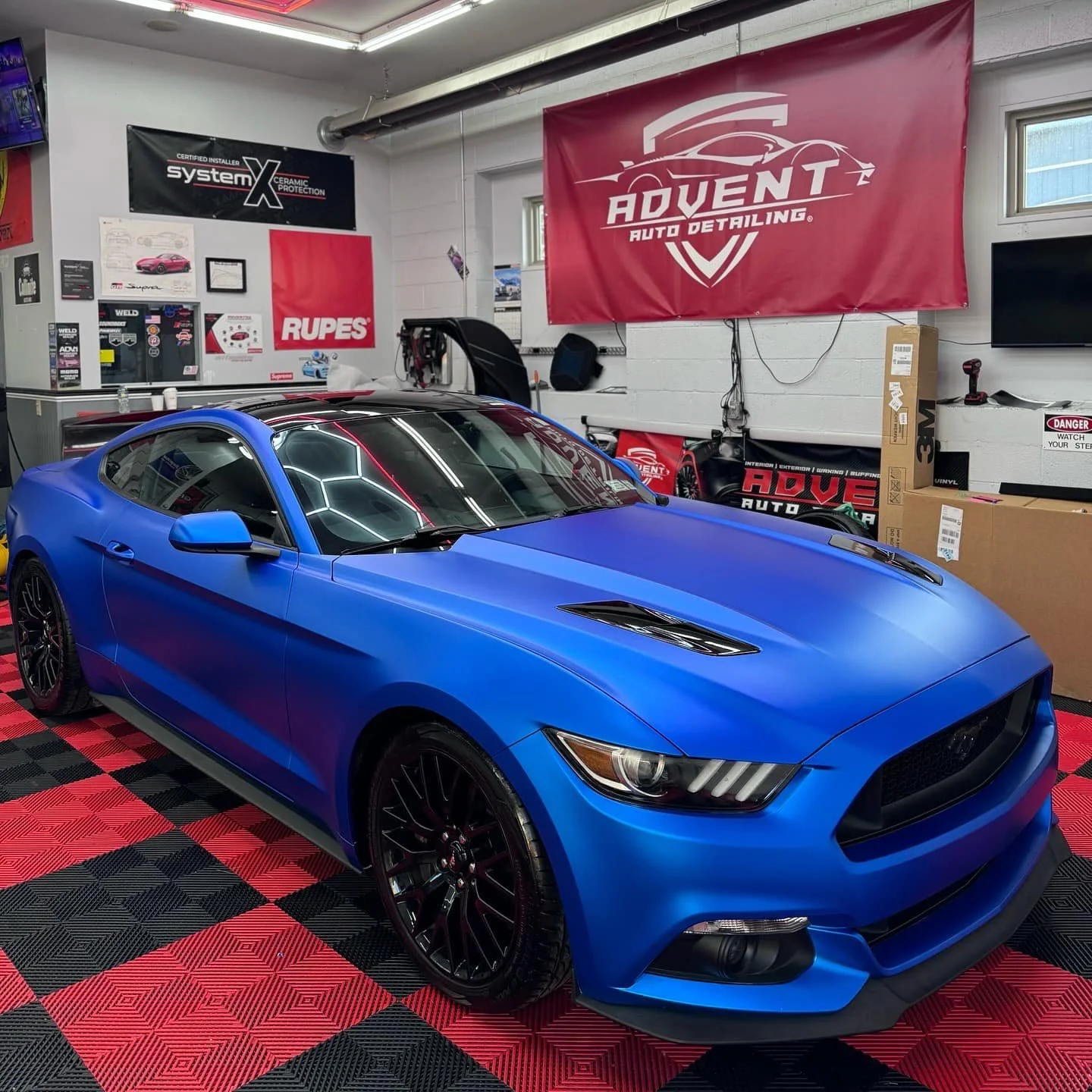 A blue Ford Mustang sports car inside an auto detailing shop with red and black checkered flooring. The shop has various banners and posters on the walls and boxes and equipment around.