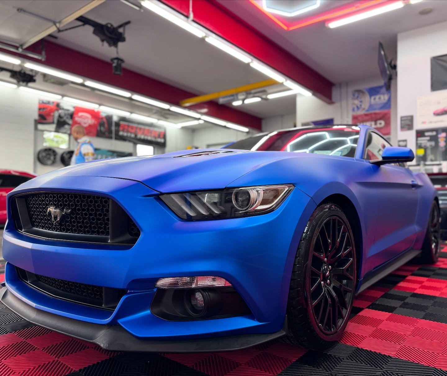A blue Ford Mustang sports car displayed inside a showroom with black wheels, low front lip, and a matte blue finish.