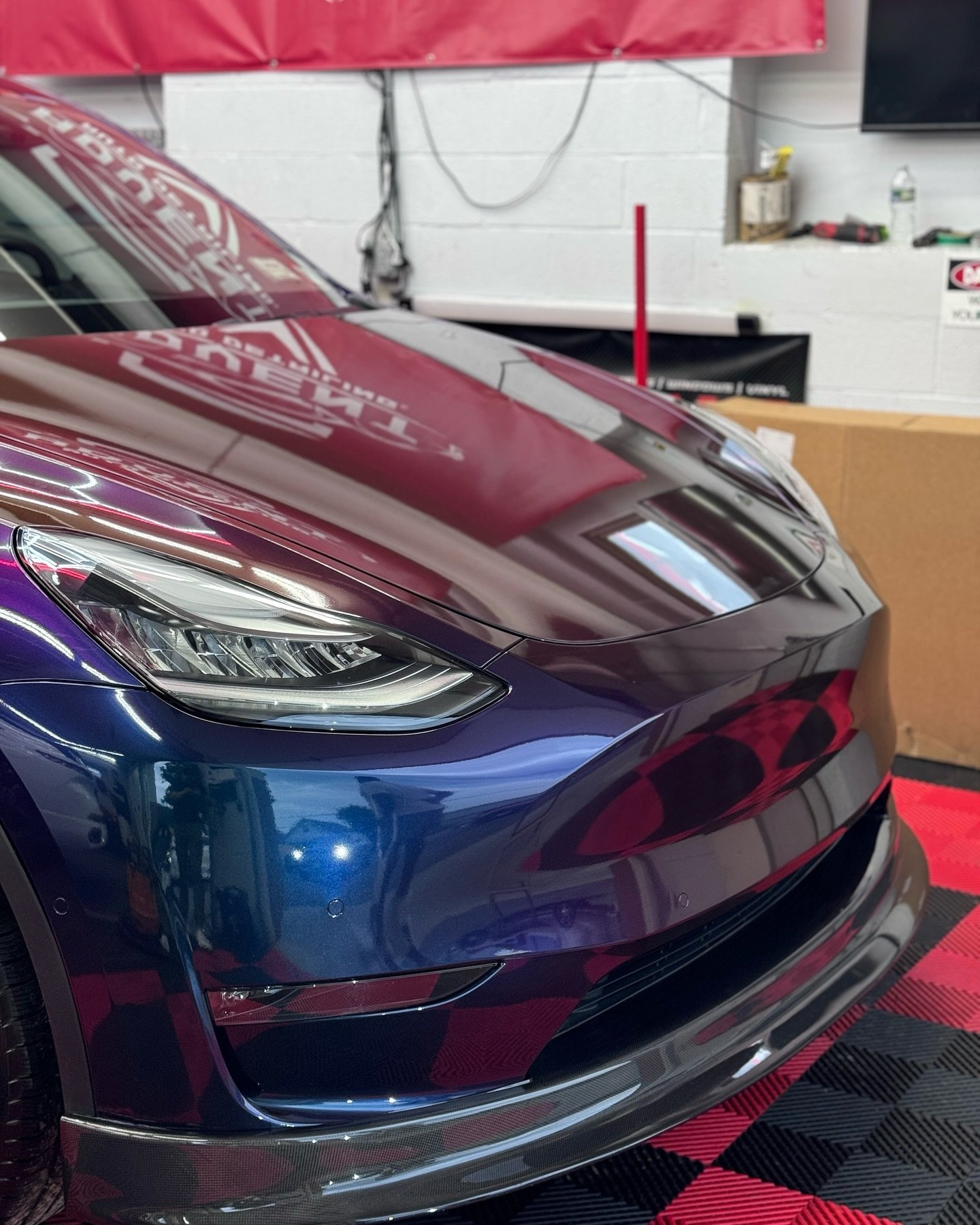 Close-up of the front of a shiny, dark blue sports car with sleek headlight design, parked indoors on a red and black checkered floor. The garage or workshop has a white brick wall, some tools, a TV, and other storage items in the background.