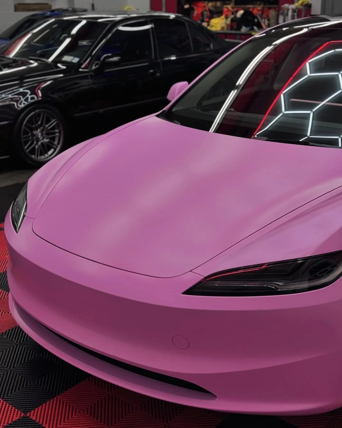 Pink sports car parked indoors, with black luxury cars in the background and neon light reflections on the windshield.
