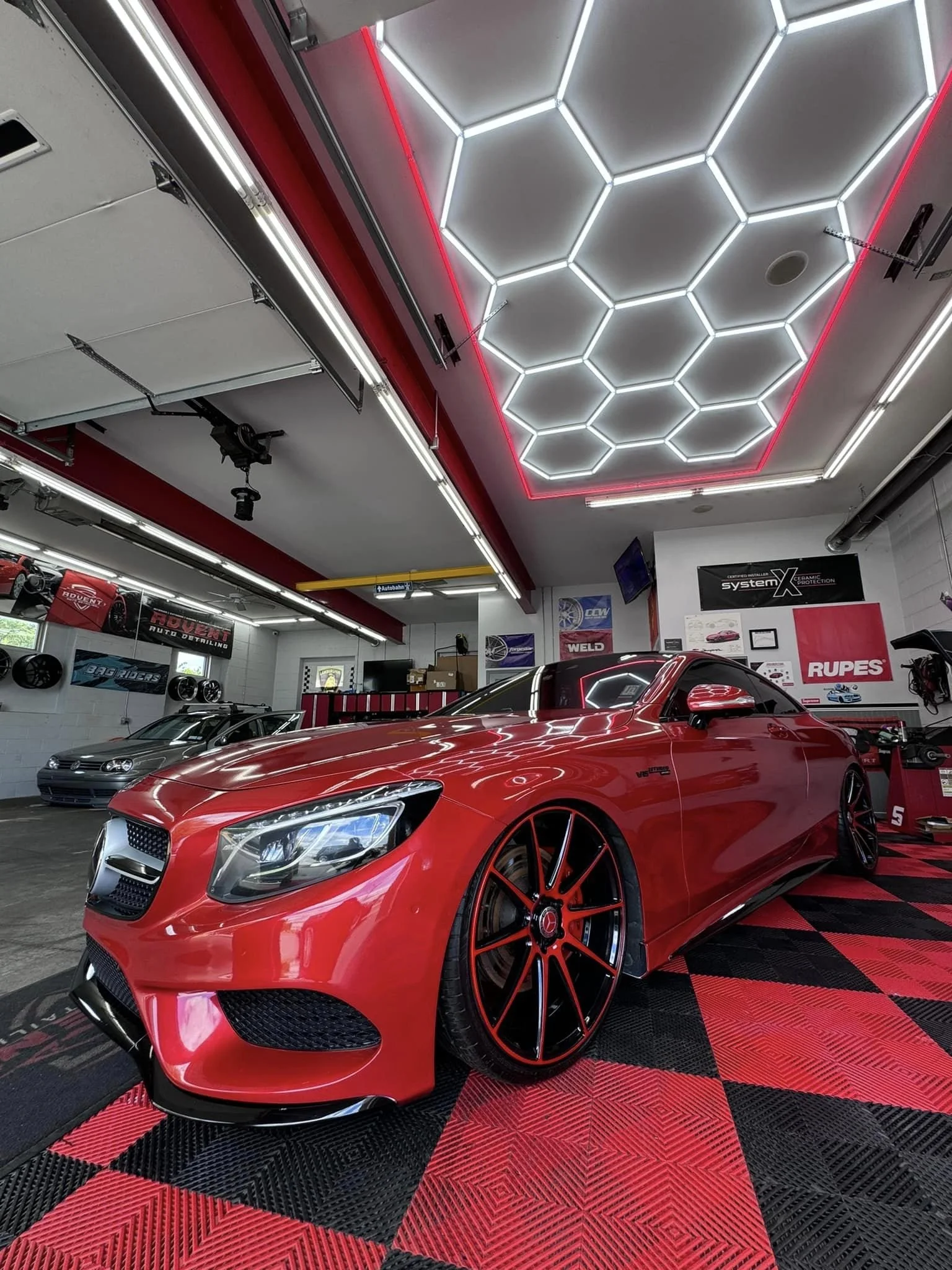Red luxury sports car parked in a garage with black and red checkered flooring, illuminated hexagon ceiling lights, and various automotive posters and equipment on the walls.