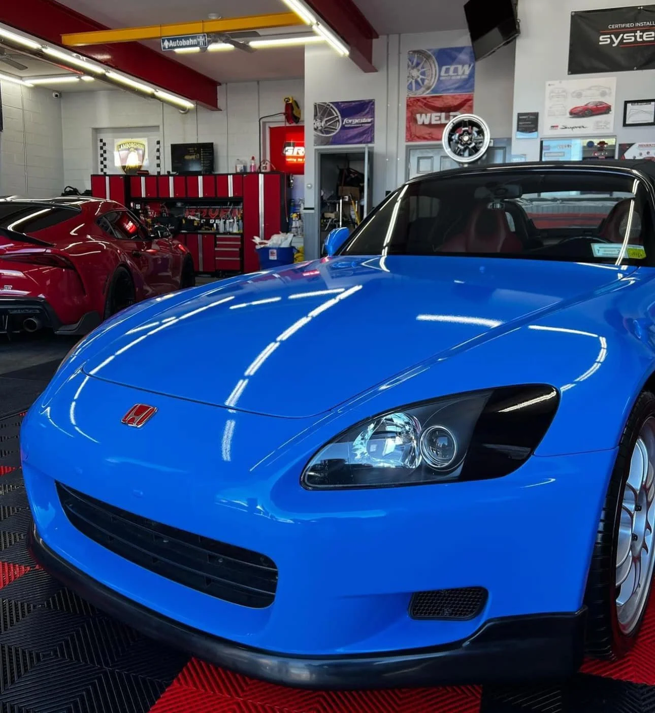 A bright blue Honda S2000 sports car parked inside a car workshop or showroom, with a red car in the background and various tools and posters on the wall.