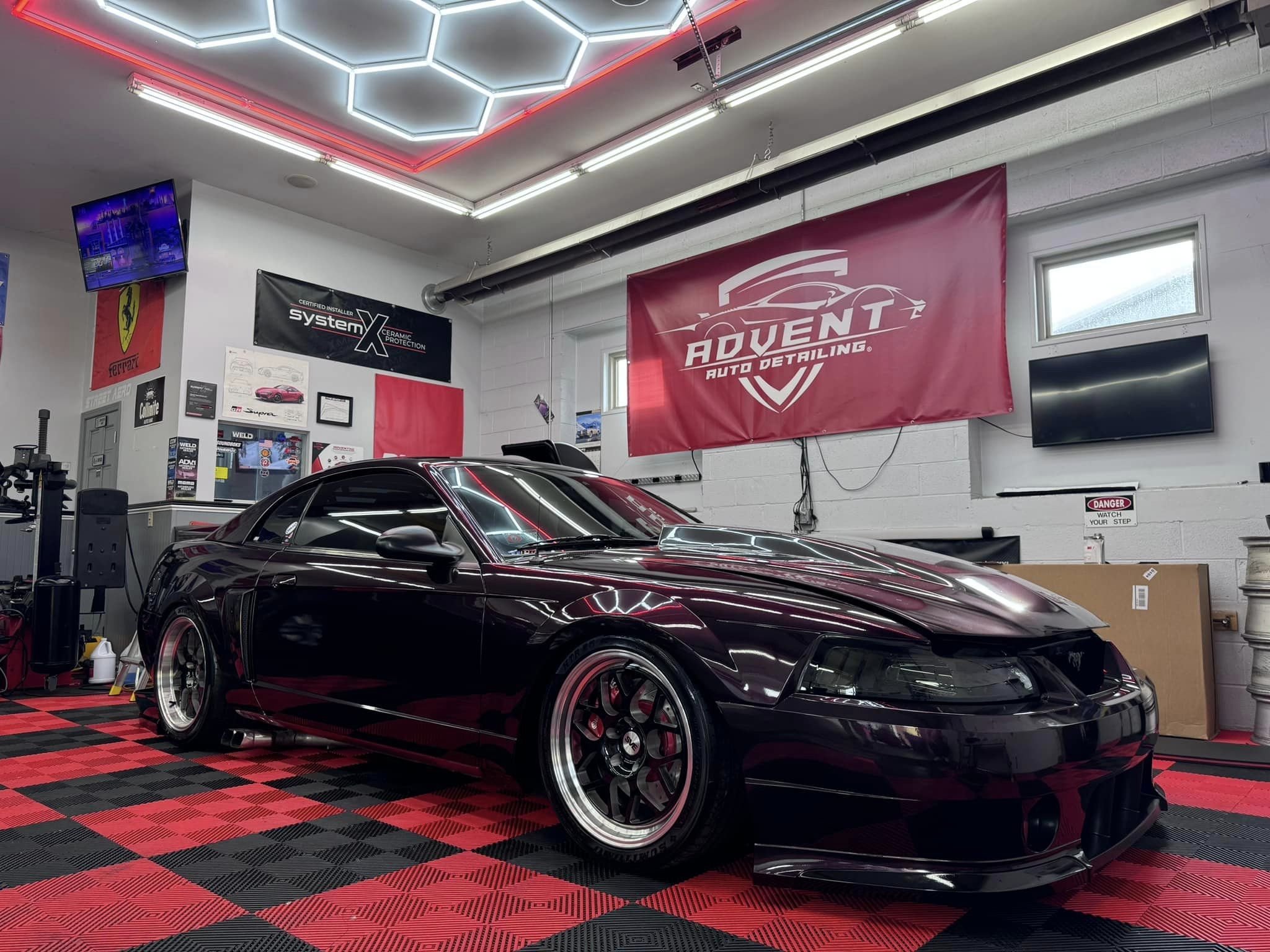 A black sports car parked inside an auto detailing shop with red and black checkered floor mats. The shop has various posters, banners, and a flat-screen TV on the walls, with a red banner reading 'Advent Auto Detailing' hanging behind the car.