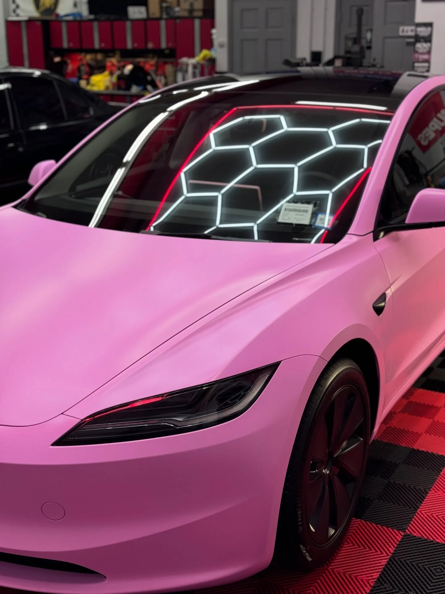A pink Tesla car inside a garage or showroom, with a honeycomb-patterned neon light display on its windshield.