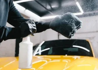 Person cleaning a yellow car with a cloth and spray bottle inside a garage
