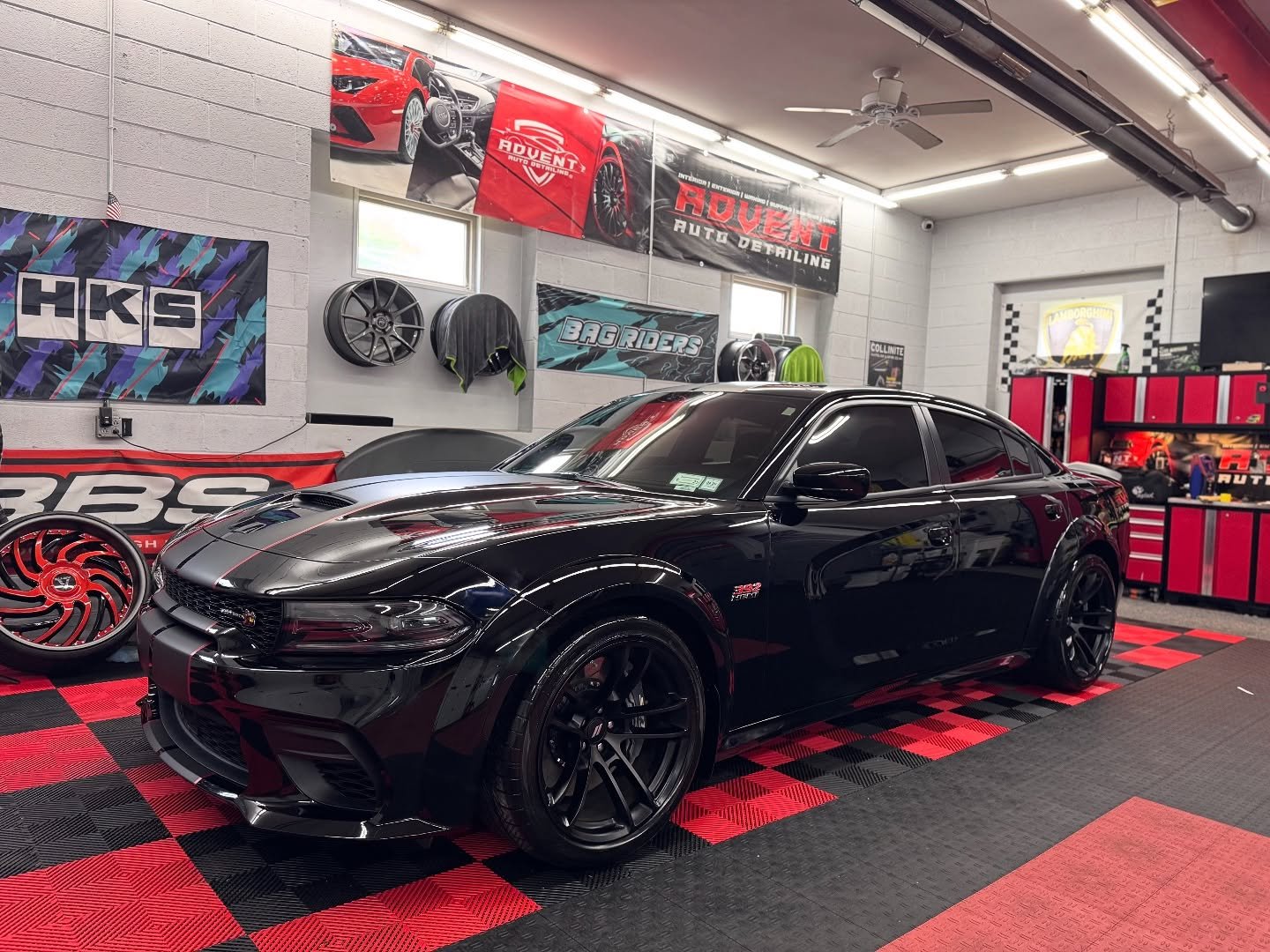 Black Dodge Charger SRT with red accents inside an auto detailing garage, surrounded by car wheels, posters, and red and black tools and storage cabinets.
