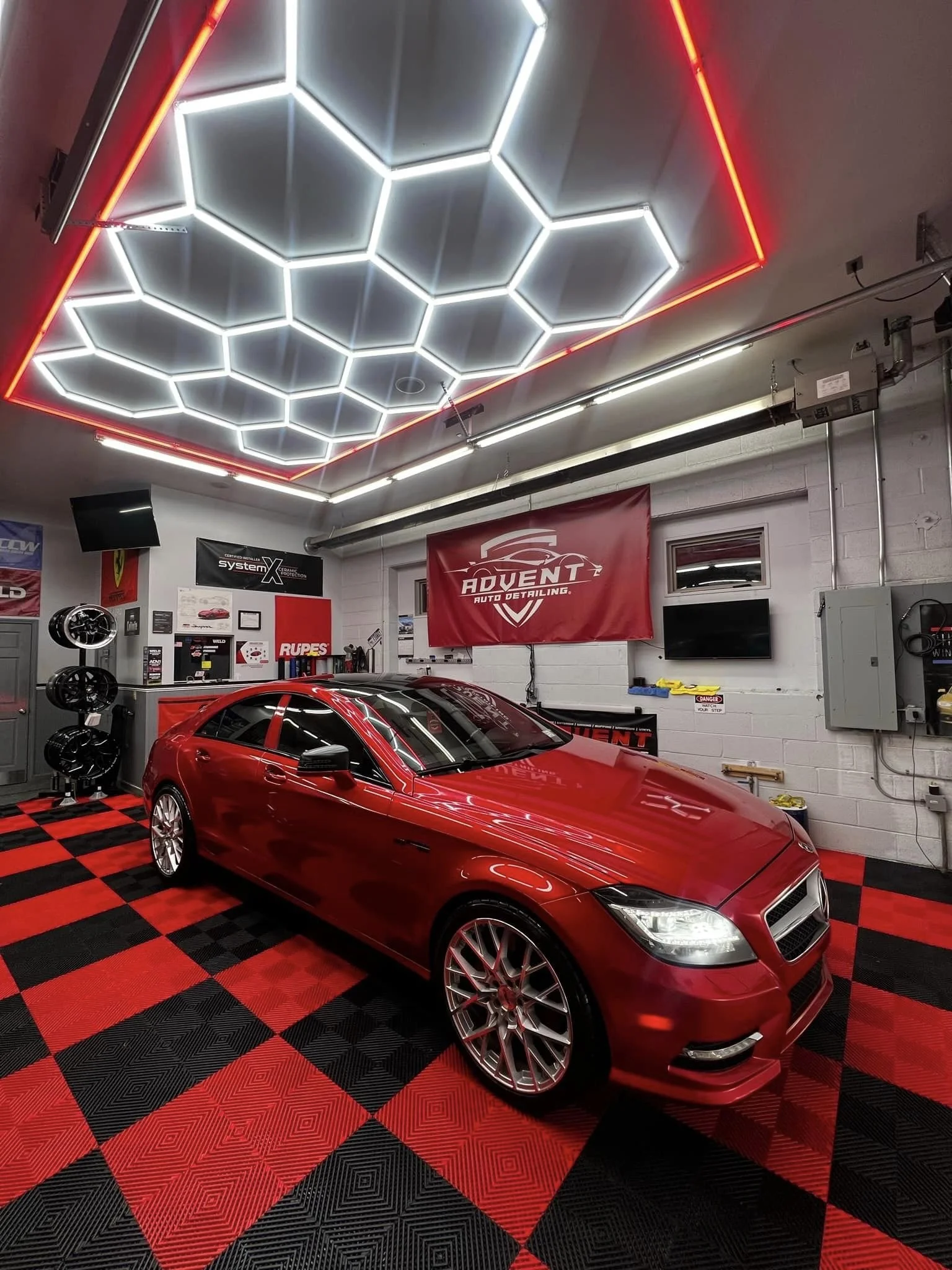 Red car inside an auto detailing garage with neon hexagon ceiling lights, black and red checkered floor, and various tools and signs on the walls.