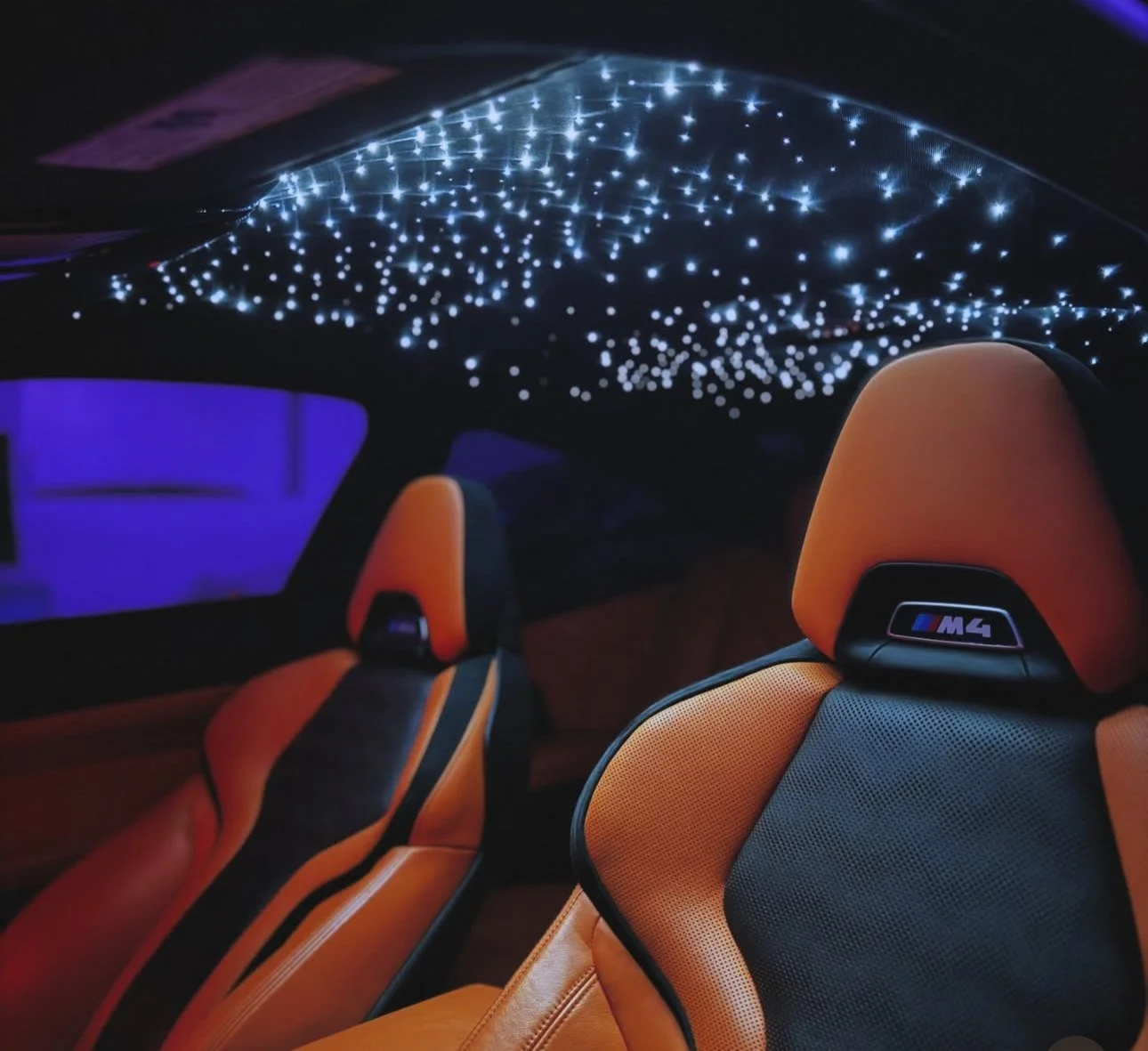 The interior of a car showing two orange and black racing seats with the BMW M4 logo, illuminated by a starry LED ceiling, with a purple light in the background.