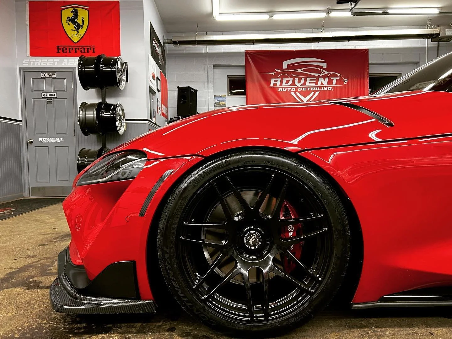 Close-up of a red sports car with black rims, parked inside an auto detailing shop. The background features a Ferrari emblem, tires on the wall, and a red banner reading "Advent Auto Detailing."