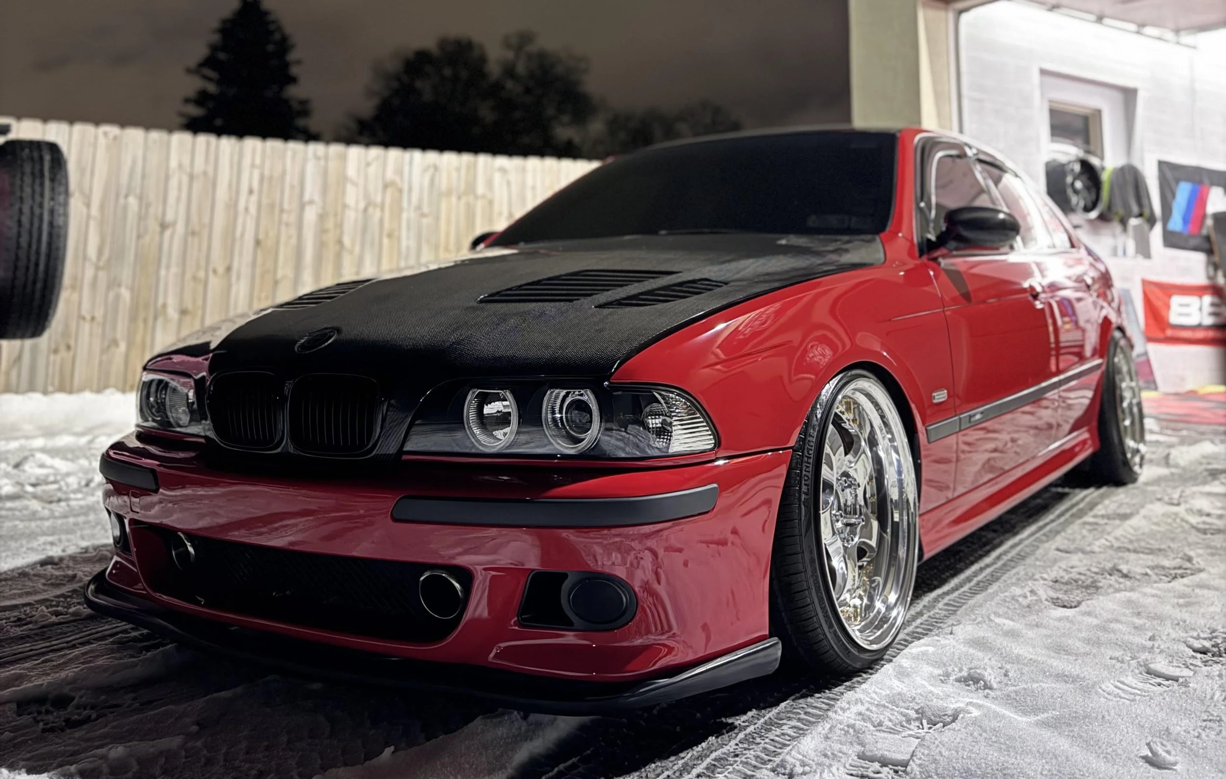 Red BMW car with black hood, parked on snow, with a wooden fence and a house in the background.