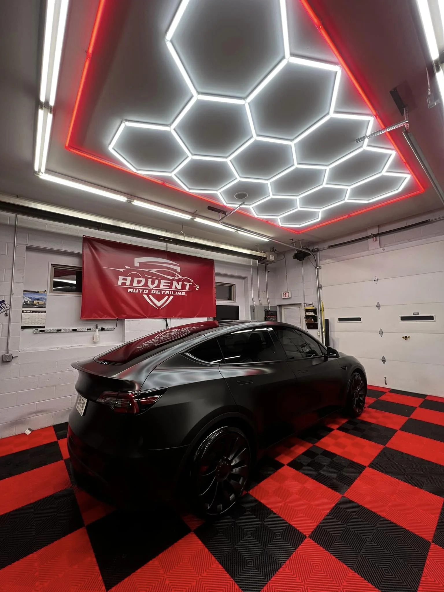 A black car parked inside a garage with red and black checkered flooring, a red banner on the wall reading 'Advent Auto Detailing,' and a ceiling with hexagonal LED light fixtures surrounded by red and white lights.