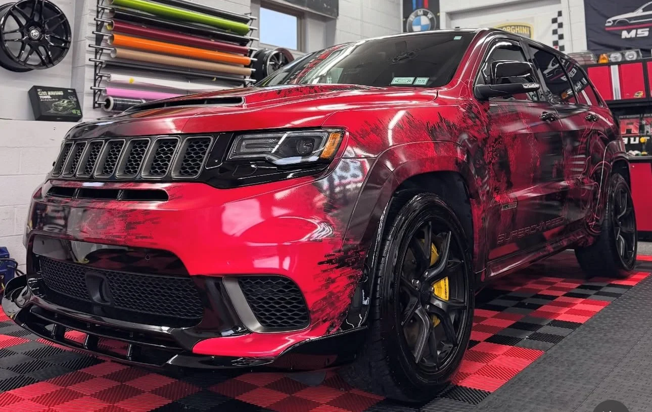 Red sports SUV with black trim and custom black wheels, inside a garage with car wrap supplies on the wall and flags in the background.