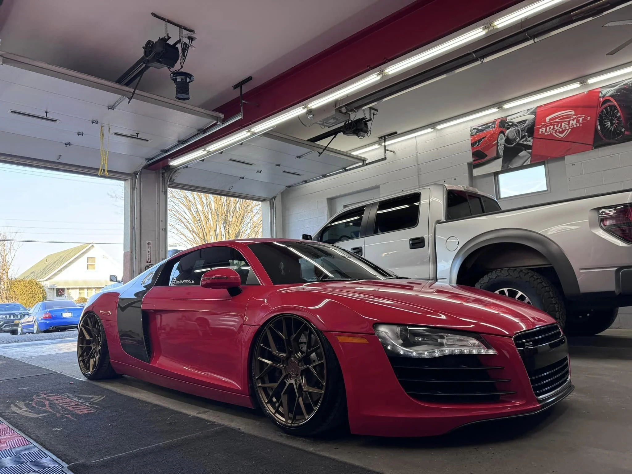 Red Audi R8 sports car and silver pickup truck inside a garage with open doors, banners on wall, and other cars outside.