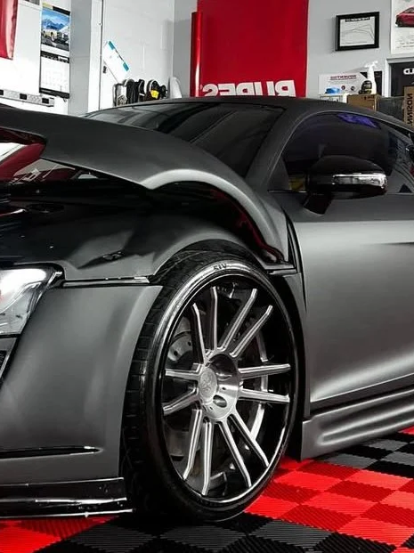 Close-up of a sleek gray sports car with an open hood, showing a modern tire and rim, in a garage with automotive posters and tools in the background.