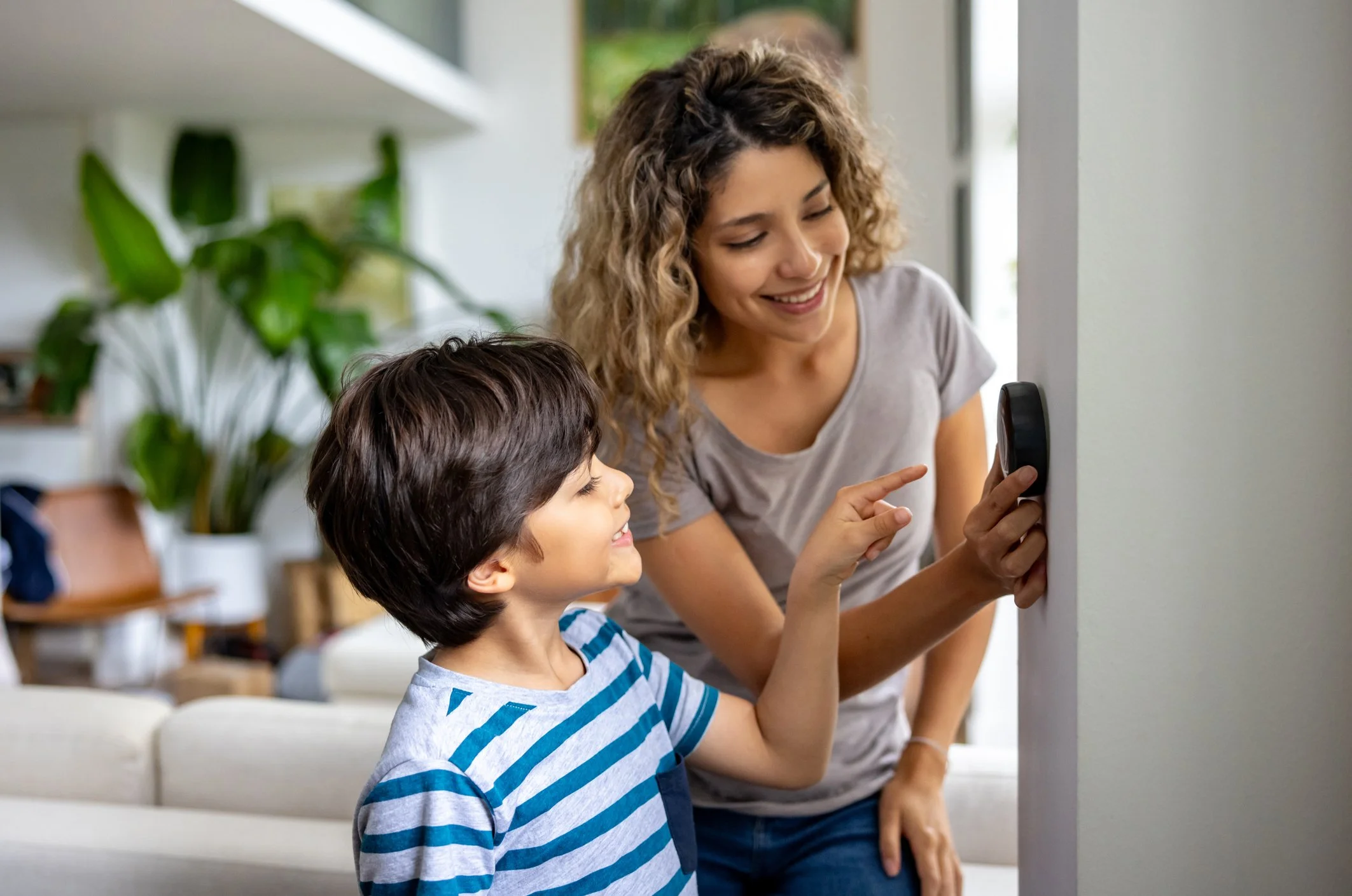 A woman and a young boy smiling and interacting with a smartphone that the woman is holding on a wall, in a bright living room with greenery and furniture in the background.