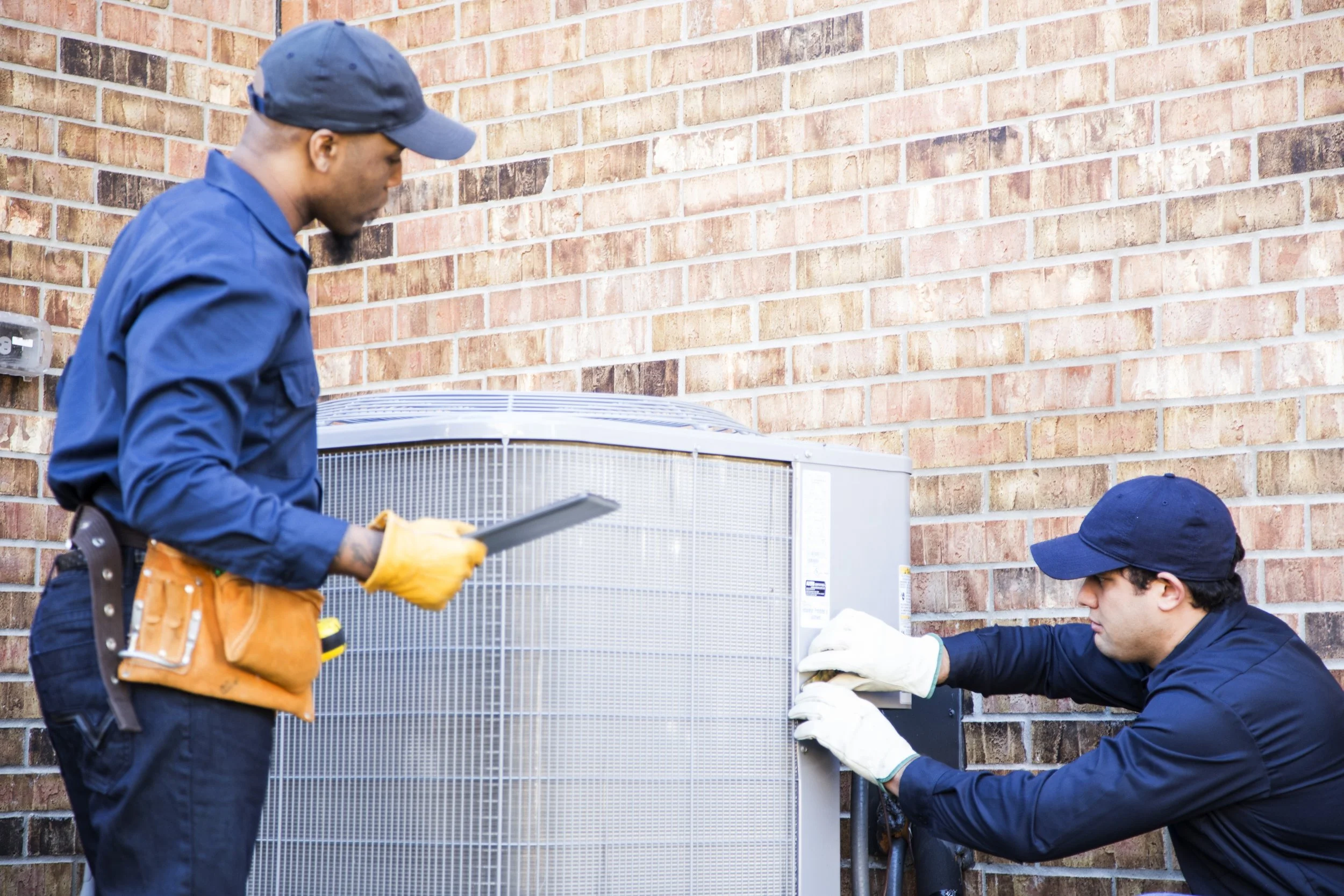 Two technicians repairing an outdoor air conditioning unit next to a brick wall, wearing gloves and work uniforms.