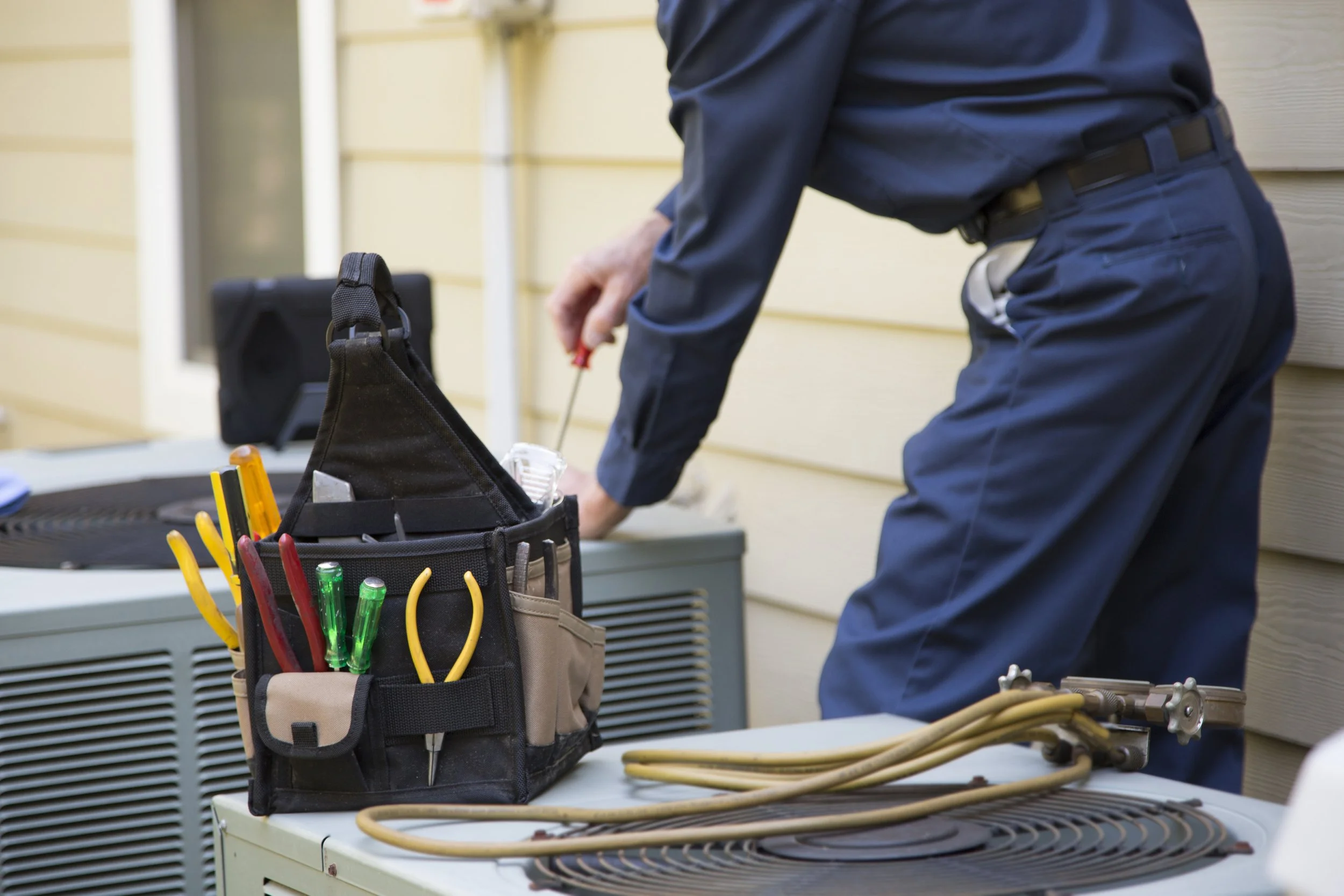 A person working on an air conditioning unit outside, with a tool bag containing screwdrivers and pliers on a table nearby.