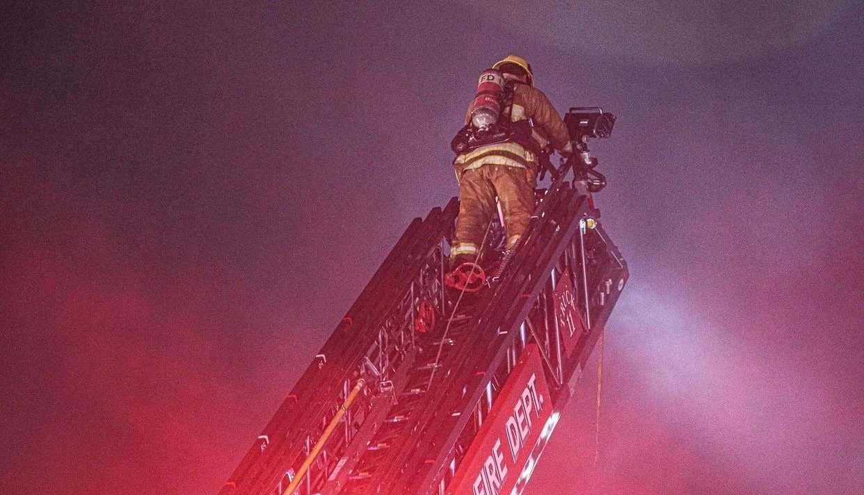 Firefighter in full gear on top of a fire truck ladder during a nighttime emergency response.