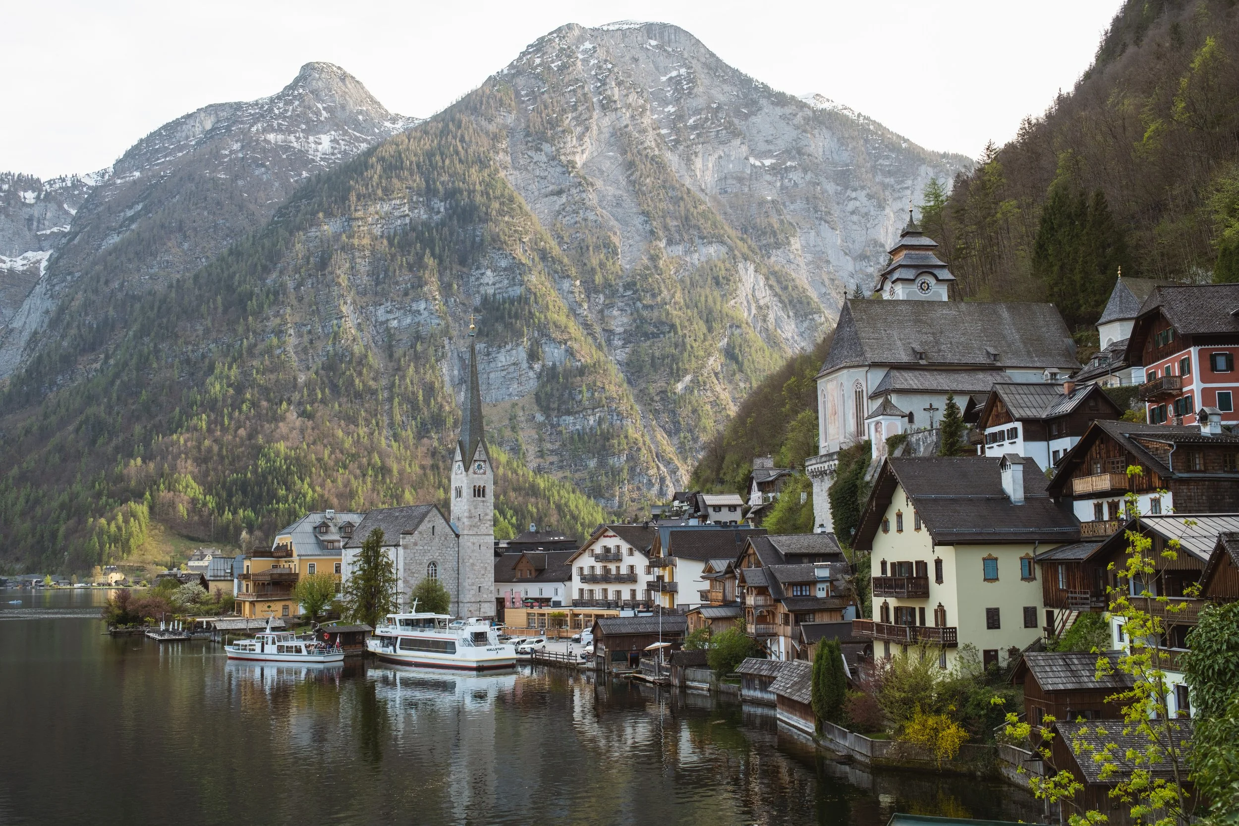 Scenic view of a small European village with colorful houses, a church, and boats on a lake, surrounded by steep, forested mountains.