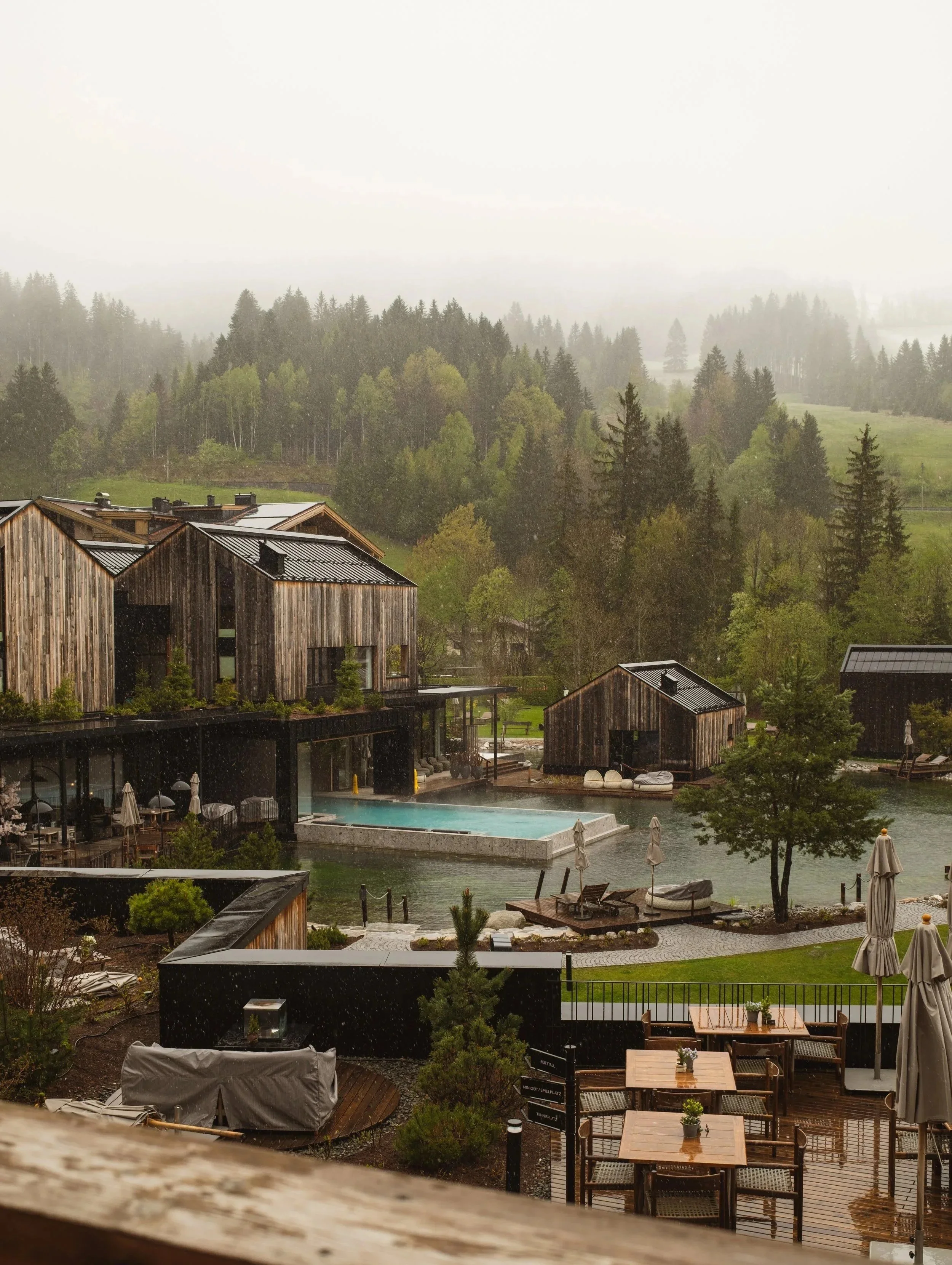 Rainy view of rustic wooden cabins with a pool, surrounded by trees and greenery, with mountains in the background.