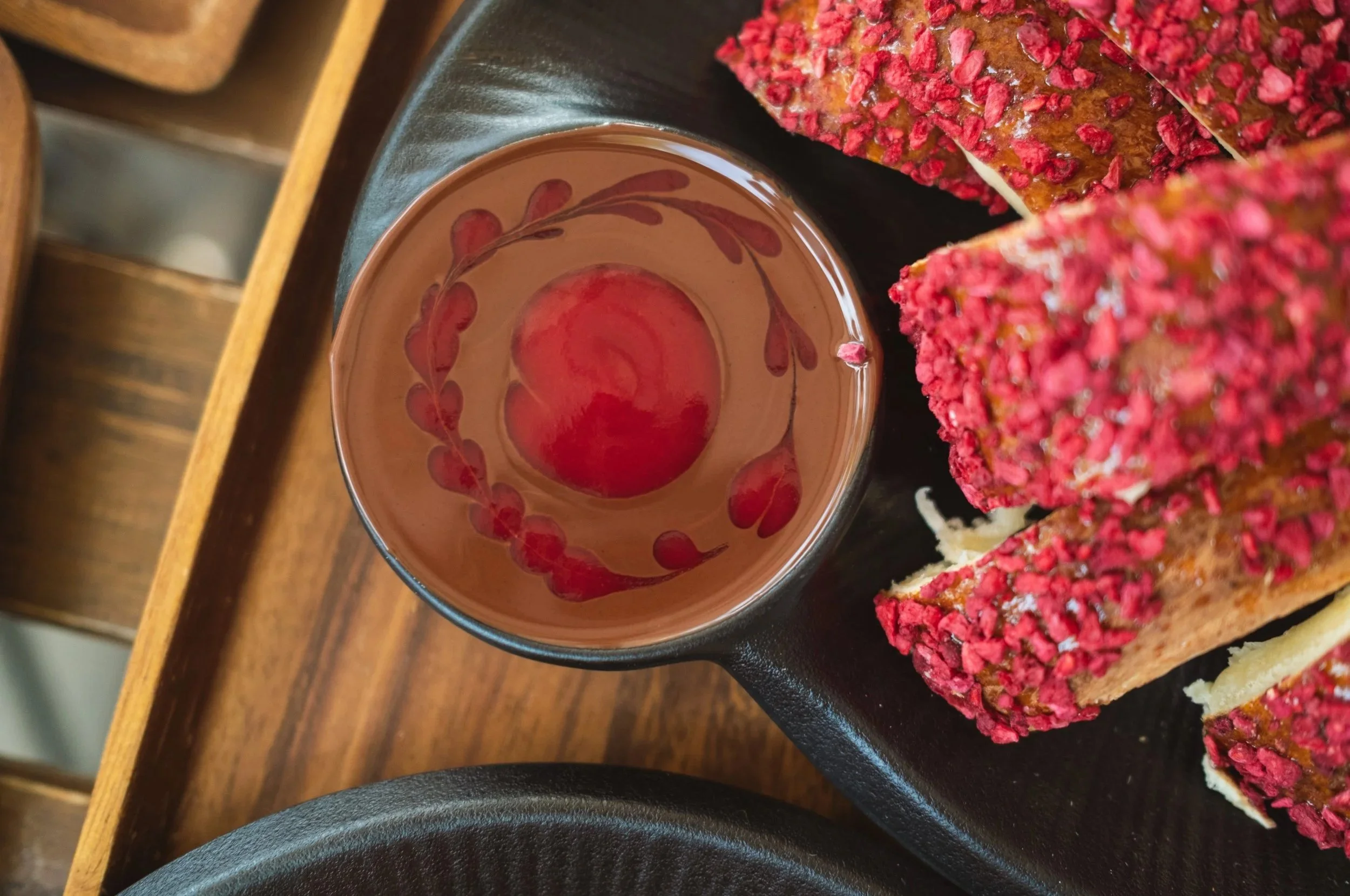 A cup of hot chocolate with a red and pink floral design on the inside, placed on a black serving tray along with pink cake slices covered in pink sprinkles, on a wooden surface.