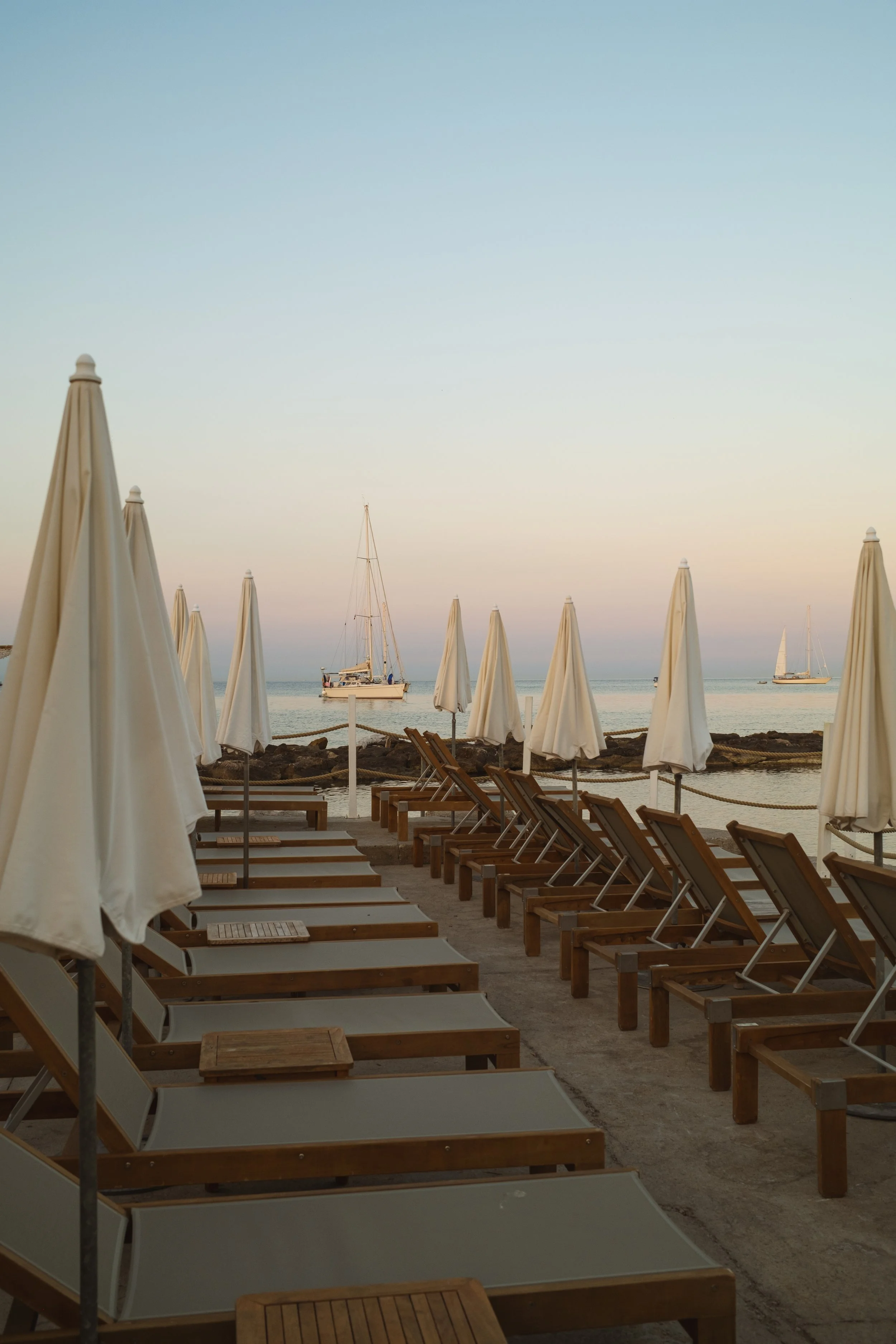 Beach scene with rows of empty lounge chairs and closed umbrellas, sailboats on calm water at sunset
