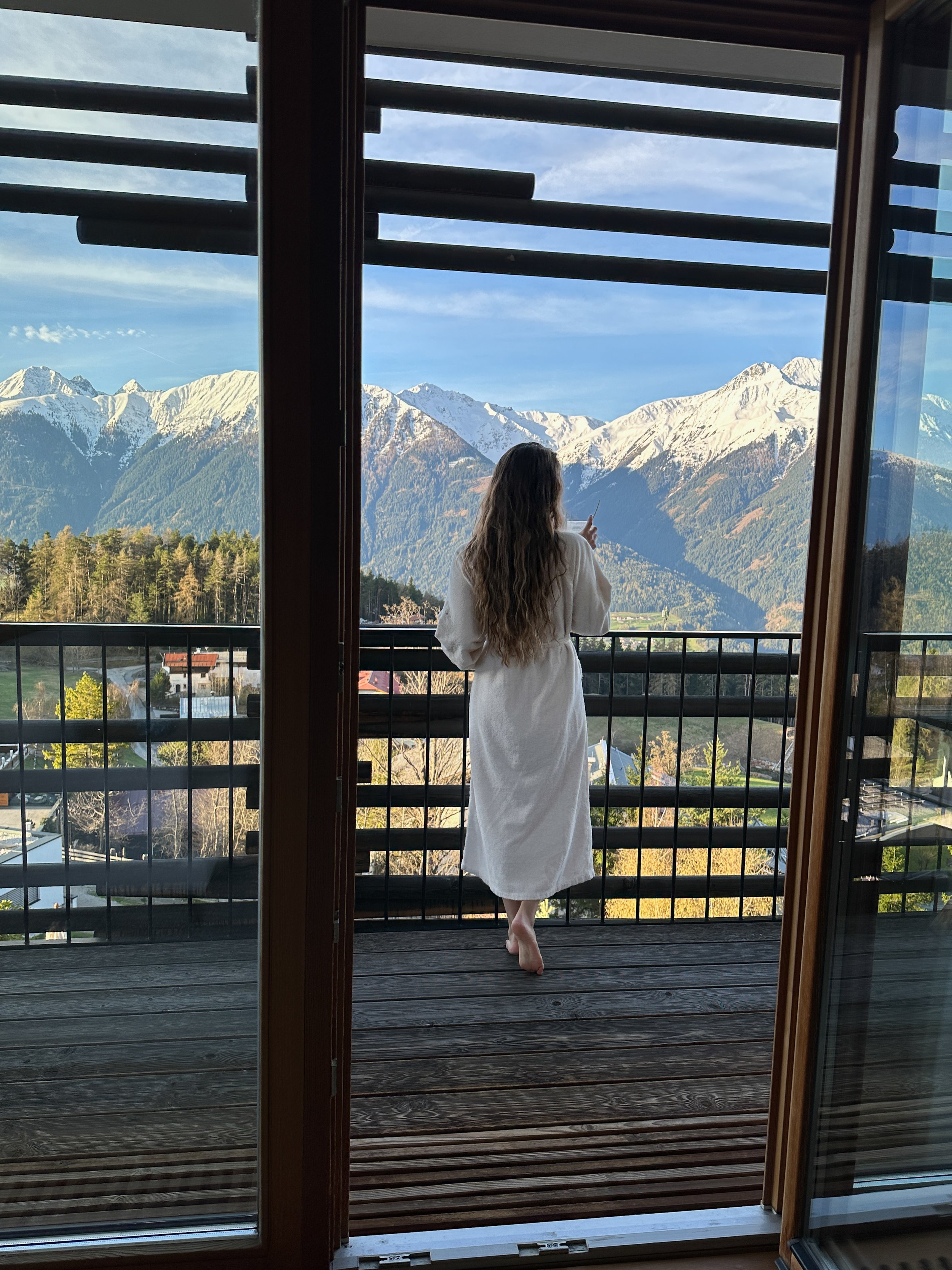 Woman in a white bathrobe standing on a balcony, looking at snow-capped mountains in the distance during daytime.