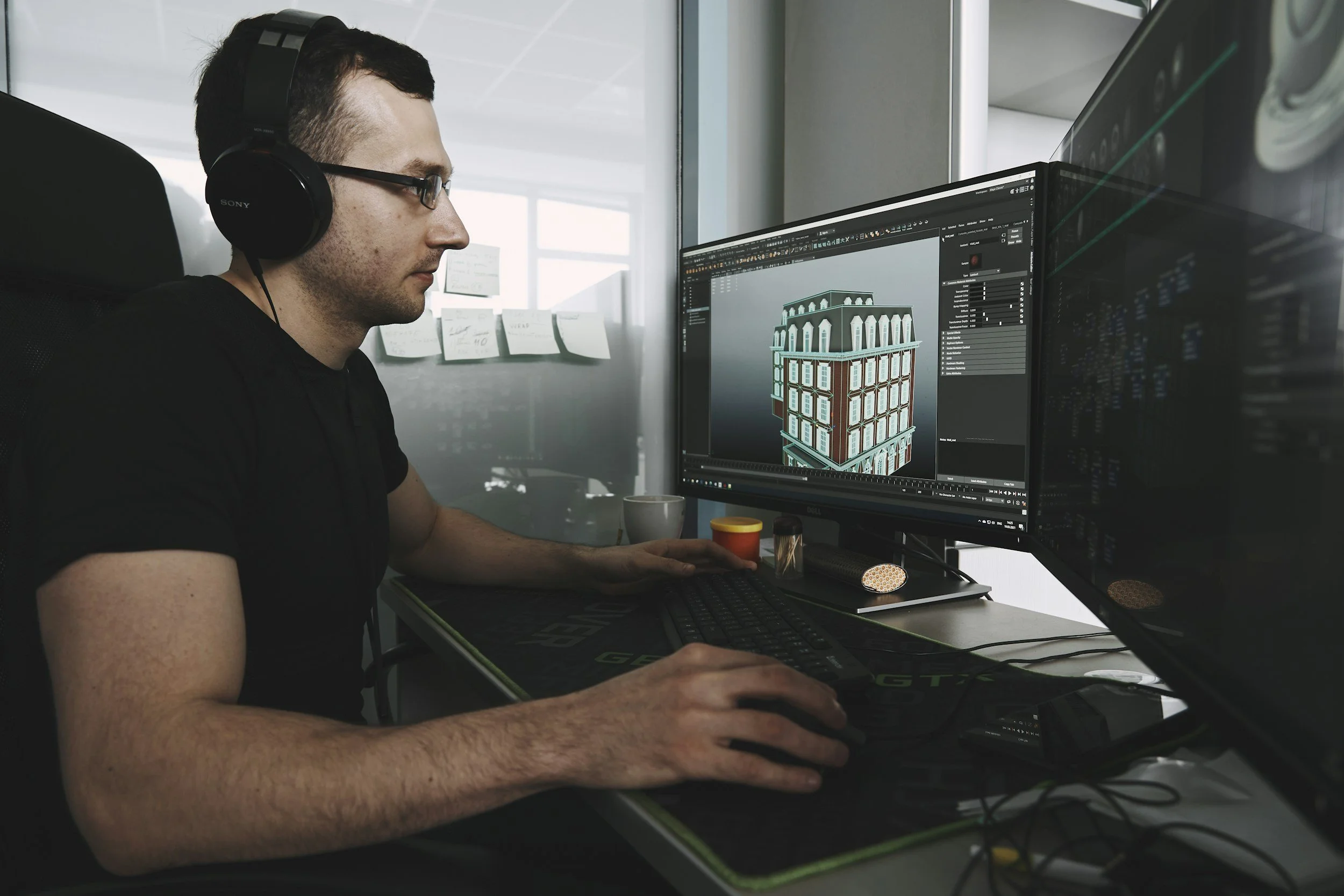 A man wearing glasses and headphones working on a computer with dual monitors, displaying a 3D architectural model of a multi-story building in a modern office.