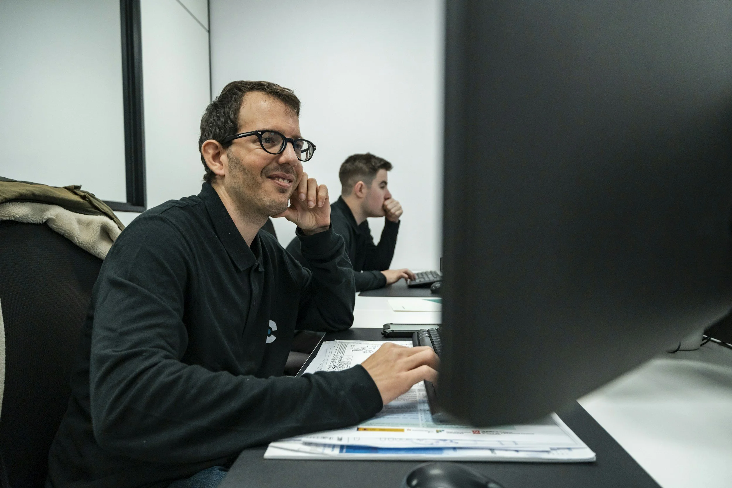 Two men working at computer desks in an office, one smiling and wearing glasses, the other focused on screen with hand on chin.