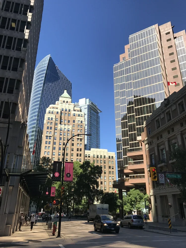 City street scene with tall modern skyscrapers, pedestrians, cars, trees, traffic lights, and street signs including Hornby Street.