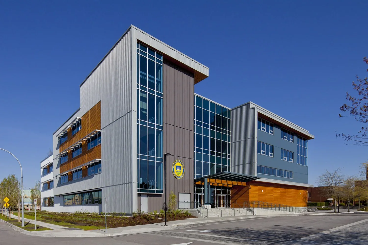 Modern multi-story building with metal siding, large glass windows, and wood accents, daytime with clear blue sky, city street in foreground.