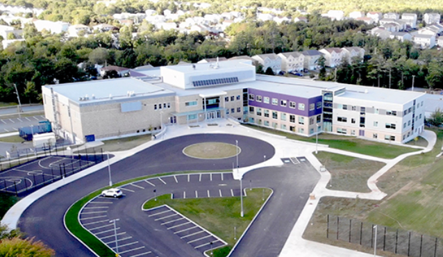 A modern school building with a parking lot, sports courts, and surrounding greenery.